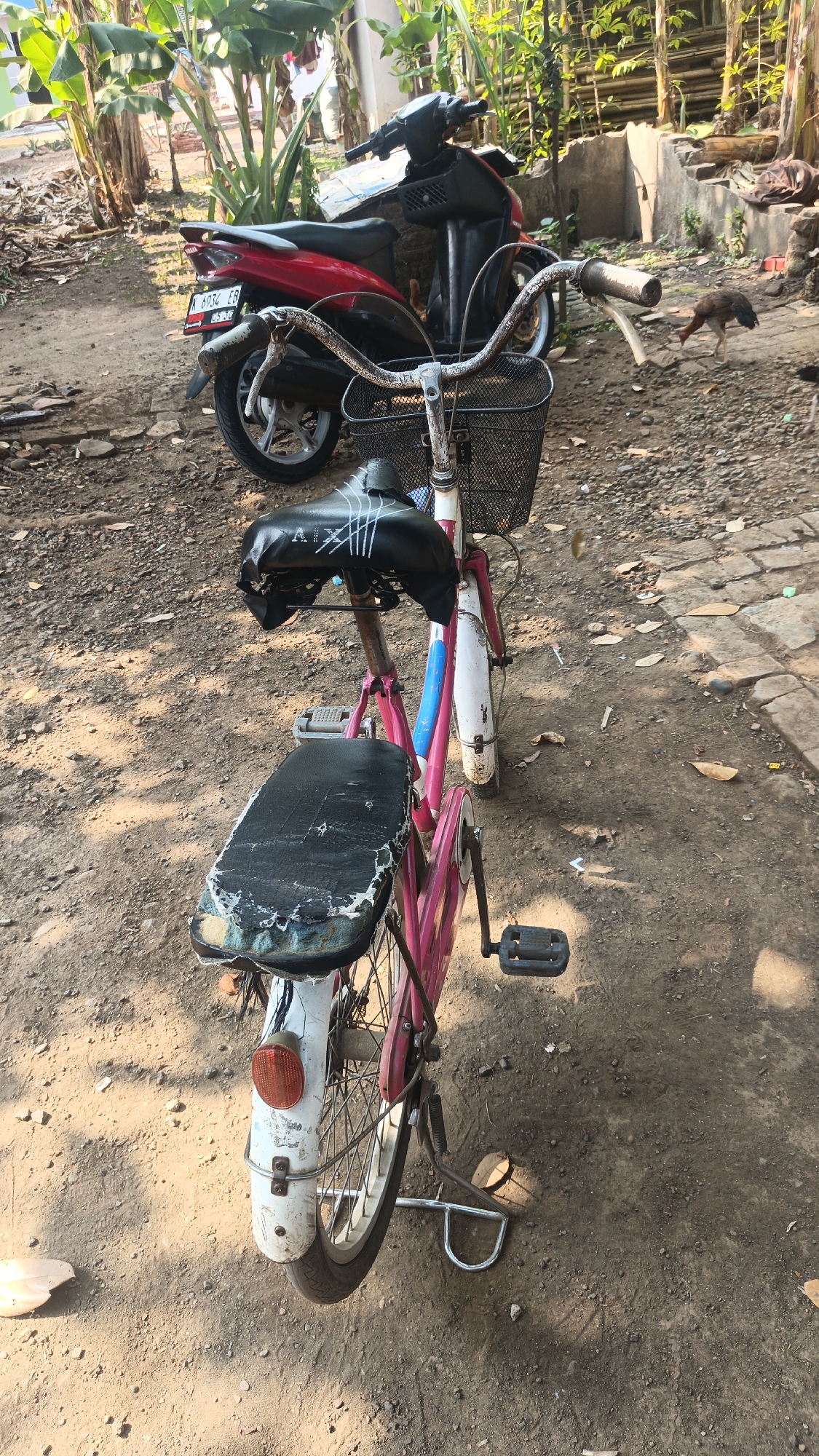 A pink bicycle with a black seat is parked on a dirt surface in what appears to be a rural or semi-rural outdoor area. A red and black scooter is parked behind the bicycle. The scene is set in Bulungcangkring, Indonesia. The ground is covered with dirt and small stones. There are some plants and trees in the background. A small dog is visible in the middle ground. The lighting suggests it is daytime. The bicycle has a basket on the handlebars and a rear rack. The seats of the bicycle show signs of wear and tear.