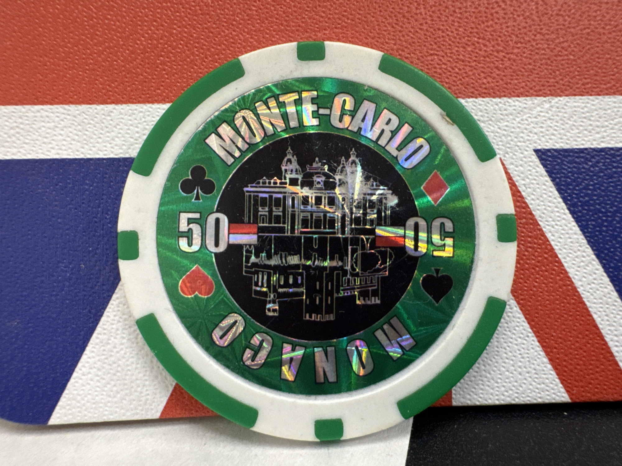 A close-up shot reveals a green and white poker chip with "MONTE-CARLO" printed on the top half and "MONACO" on the bottom. The chip displays the value "50" on both sides of a building graphic in the center. The suits of cards (clubs, diamonds, hearts, spades) are placed between the text and building. The chip lies on a surface with a pattern of blue, white, and red stripes, possibly a flag design. There is no indication of Yerevan, Armenia.