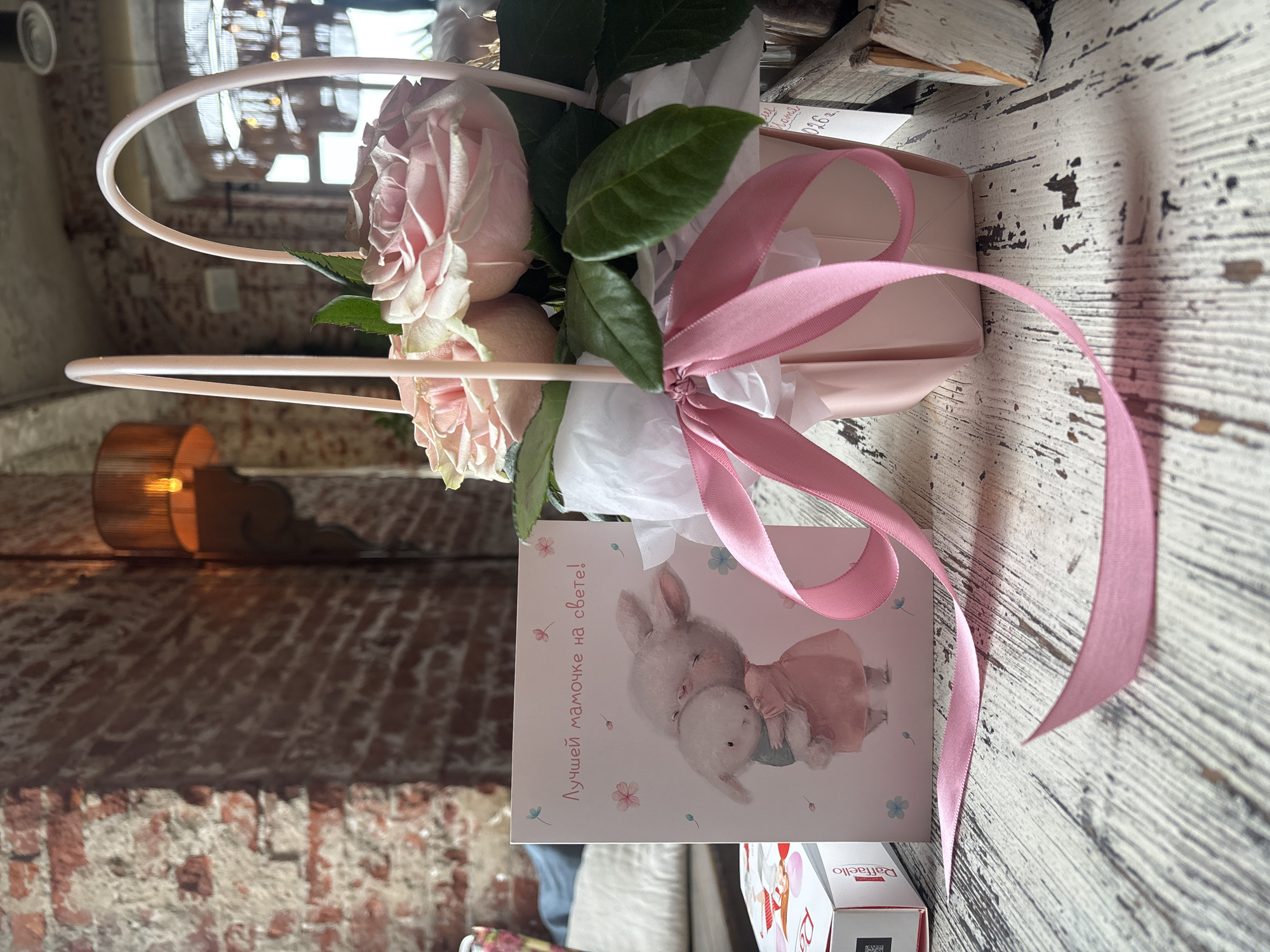 A close-up shot shows a pink flower arrangement in a light pink basket, accompanied by a greeting card and a box of chocolates. The card features an illustration of two rabbits hugging, with the Russian text "Лучшей мамочке на свете!" (To the best mom in the world!). The flowers appear to be light pink roses. A pink ribbon is tied around the basket. To the left, a white box with red accents, identified as "Raffaello," is partially visible. The items are arranged on a rustic wooden surface with a distressed white finish. In the background, a brick wall and a warm light fixture are visible, suggesting an indoor setting, possibly a cafe or restaurant. No specific location in Ivanovo, Russia, can be determined from the visual information.
