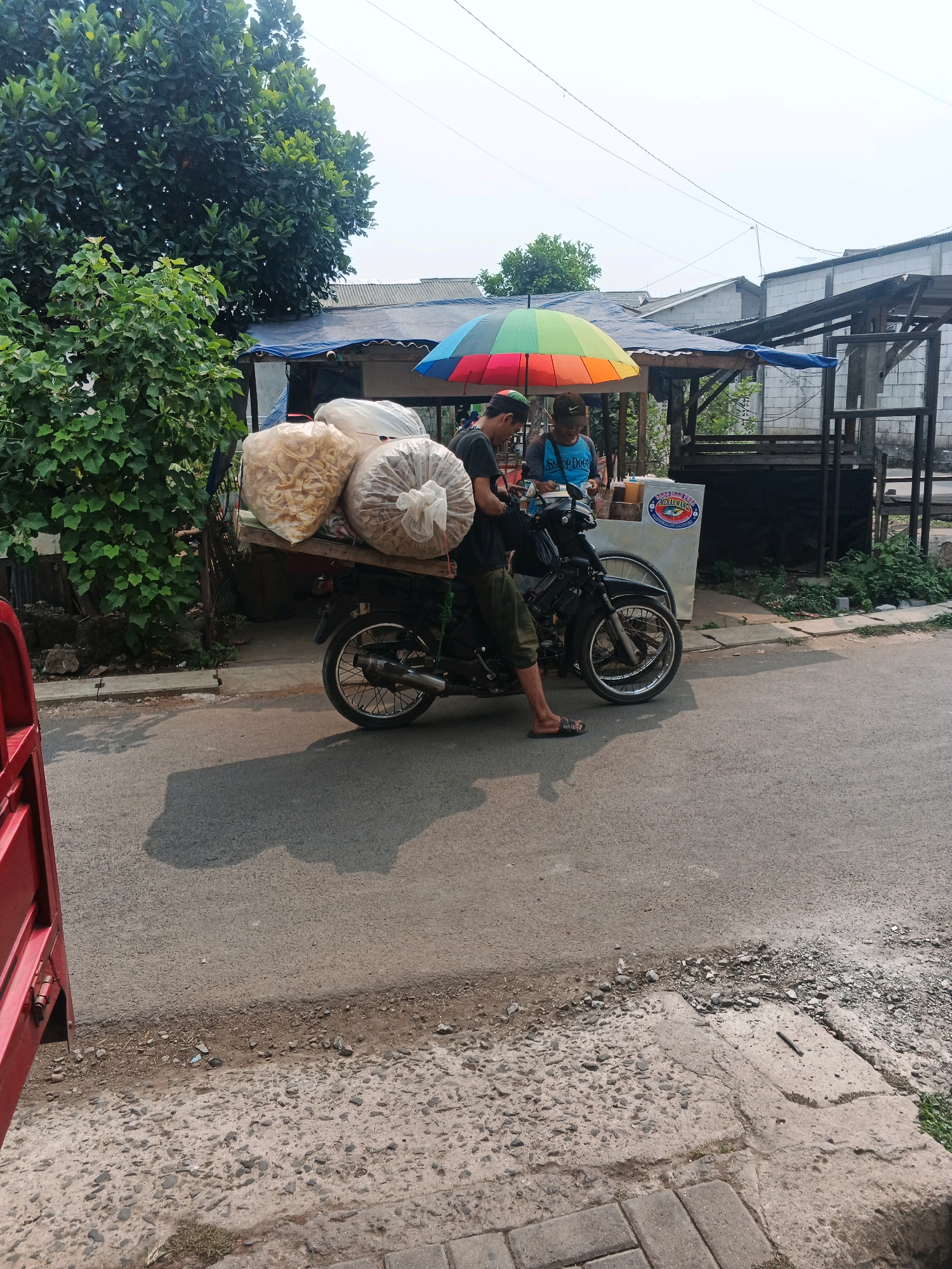 A man wearing a black t-shirt and olive green pants stands next to a motorcycle loaded with two large sacks of snacks. The motorcycle is parked on an asphalt road in front of a small food stall. The stall has a blue tarp roof and a counter displaying various food items. A rainbow-colored umbrella is set up over the stall, and two young boys are visible behind the counter. A large tree with dense green foliage is to the left of the scene, and buildings are visible in the background. The setting appears to be a street in Kadu, Indonesia. The man is likely a vendor preparing to transport or sell the snacks, and the boys are likely assisting him at the food stall.