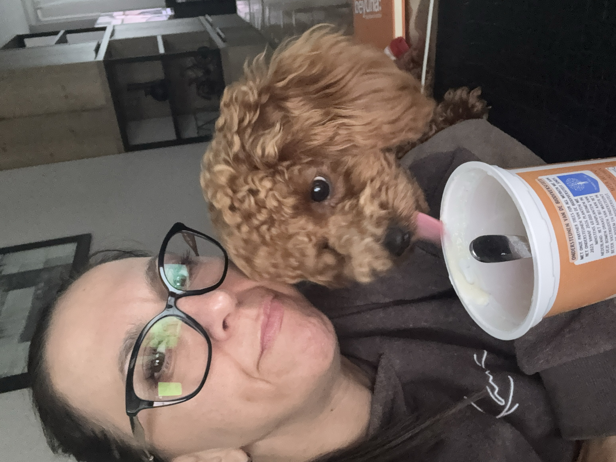 This is a selfie taken by a woman with glasses, lying down on what appears to be a couch. A small, reddish-brown poodle is affectionately leaning its head onto the woman's face, with its tongue extended towards a container of "beyuna" yogurt that the woman is holding. The dog's eyes are wide and appear to be looking at the camera. The woman is smiling slightly, and the overall mood is one of warmth and affection. The background shows shelving units that suggest an indoor setting, possibly a living room. Text visible on the yogurt container reads "ONDERSTEUNEN VAN DE BIODIVERSITEIT" which translates to "SUPPORTING BIODIVERSITY." This indicates that the product is involved in an environmental initiative, with Trees for All planting trees to support biodiversity. The image appears to be taken indoors during the daytime.