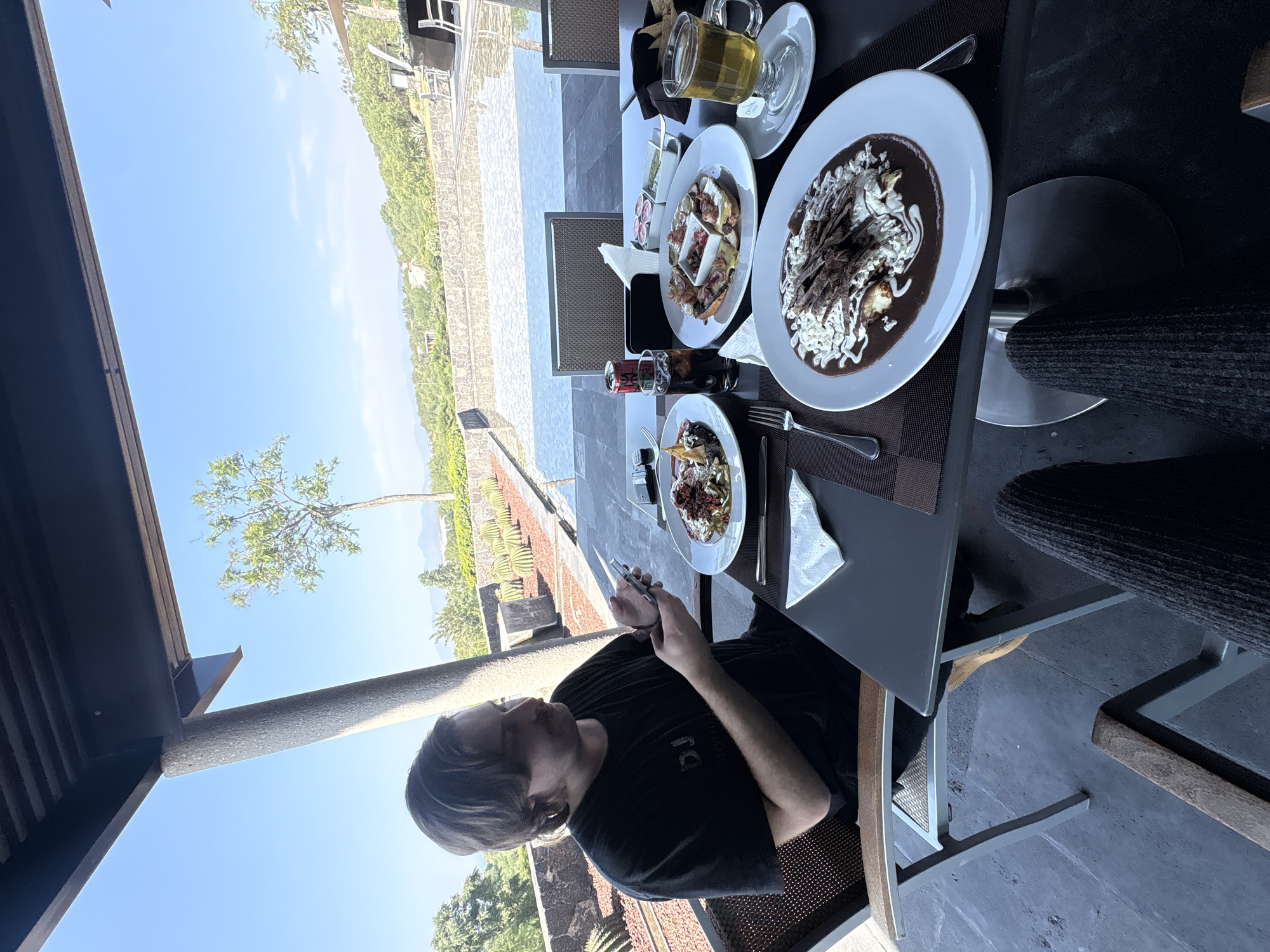 A man sits at an outdoor table in Pueblo Nuevo de Jasso, Mexico, looking at his phone. The table is set with plates of food, including what appears to be enchiladas, drinks, and condiments. The background shows a sunny day and a landscape of trees and buildings.