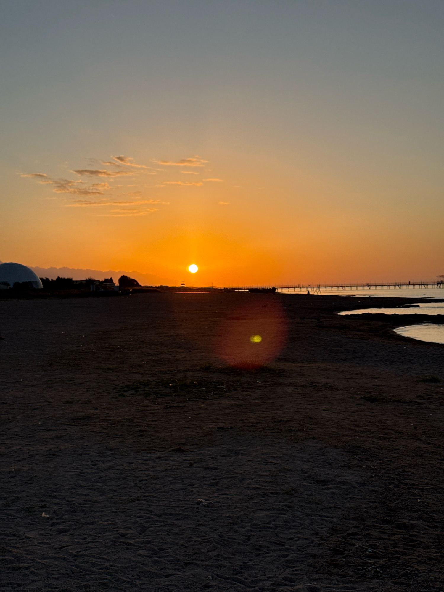 The image depicts a sunset at Lake Issyk-Kul in Bosteri, Kyrgyzstan. The sun is low on the horizon, casting an orange glow across the sky and the calm water. A long wooden pier extends into the lake, with silhouetted figures of people visible on it. The foreground is a dark, sandy beach, with some scattered debris. In the distance, a large, domed structure is partially visible on the left, and low-lying hills form the backdrop. The overall scene is serene and peaceful, with the warm colors of the sunset dominating the image.