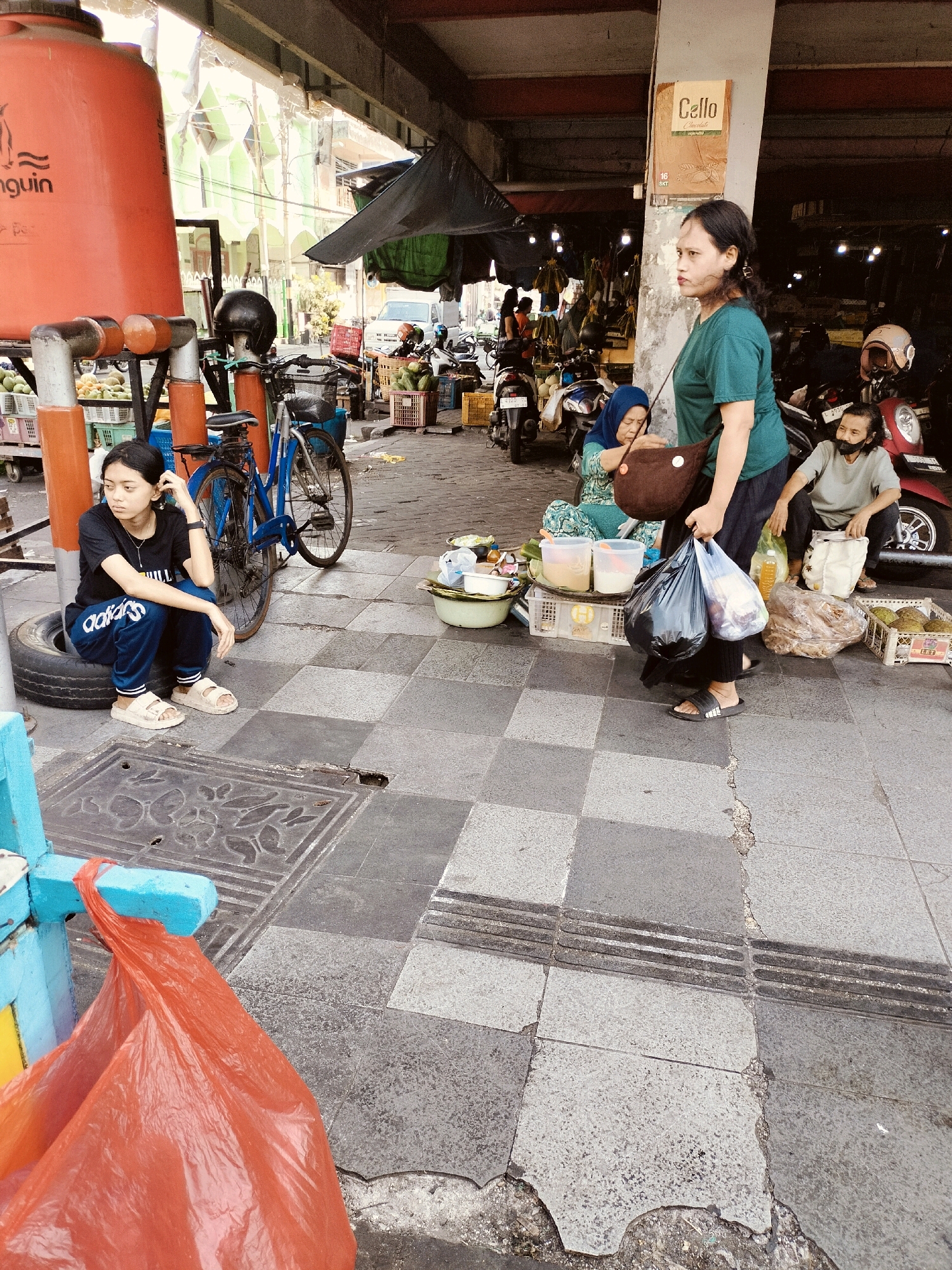 In an outdoor market setting in Surabaya, Indonesia, a young woman in a black t-shirt and blue Adidas pants sits on a tire, looking to her left. A blue bicycle is parked behind her. To her right, a woman wearing a blue hijab and a patterned green dress is interacting with a woman in a green t-shirt and dark pants. The latter is holding two black plastic bags filled with groceries. A man wearing a grey t-shirt and a black mask sits further to the right, near a red motorcycle. The ground is paved with grey and black checkered tiles, some of which are cracked and damaged. Various food items are displayed on crates and in bowls, suggesting a marketplace or food stalls. A large orange water tank is visible on the left side of the image, along with a red plastic bag hanging from a blue structure in the foreground. The background reveals more market stalls, motorcycles, and buildings, indicating a bustling urban environment.