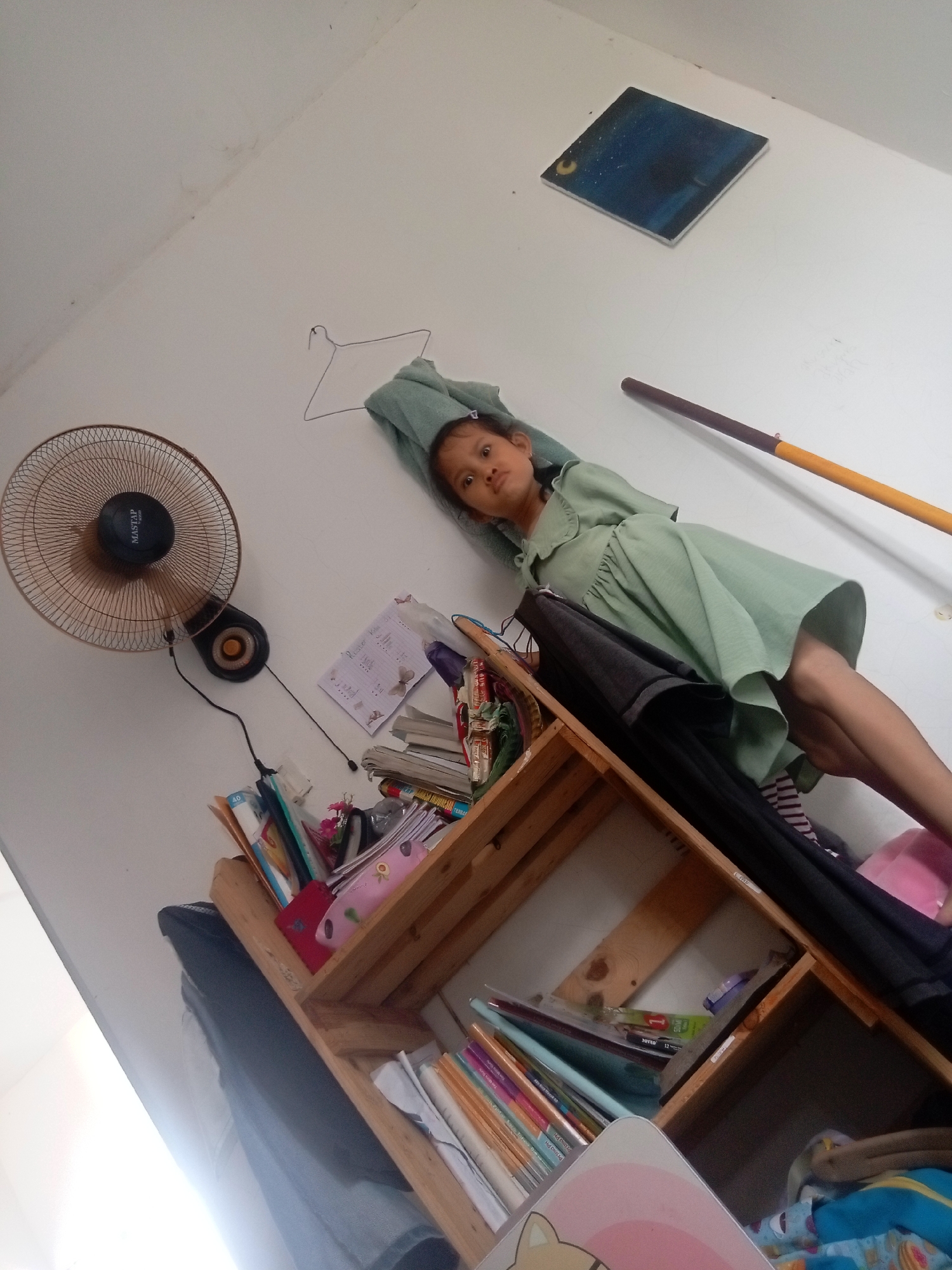 In this medium shot, a young girl, possibly around five years old, is the central subject. She is lying on her back against a white wall, her head resting on a light green towel draped over a clothes hanger. She wears a light green dress with a ruffled collar and a gathered waist. Her eyes are open and looking towards the camera with a neutral expression.
The setting appears to be an indoor space, likely a bedroom or play area, with a bright, possibly daytime, light source casting illumination from the left side of the frame, creating a strong contrast with the darker shadows. To the left of the girl, a round, brown electric fan is mounted on the wall. Below the fan, a wooden shelf unit is filled with various items. These include books, notebooks, pens, and what appear to be art supplies. Some of the books have visible spines with colorful text. A few small pieces of paper with writing are also scattered on the shelves.
On the wall above the girl, a small, square painting depicts a dark blue night sky with stars and a crescent moon. To the right of the painting, a broom or mop with a wooden handle and a yellow tip is leaning against the wall. The overall impression is a casual, everyday scene, perhaps a moment of rest or observation in a child's room. The location cues suggest Batam, Indonesia. No specific time of day or weather is discernible, beyond the implied interior lighting.