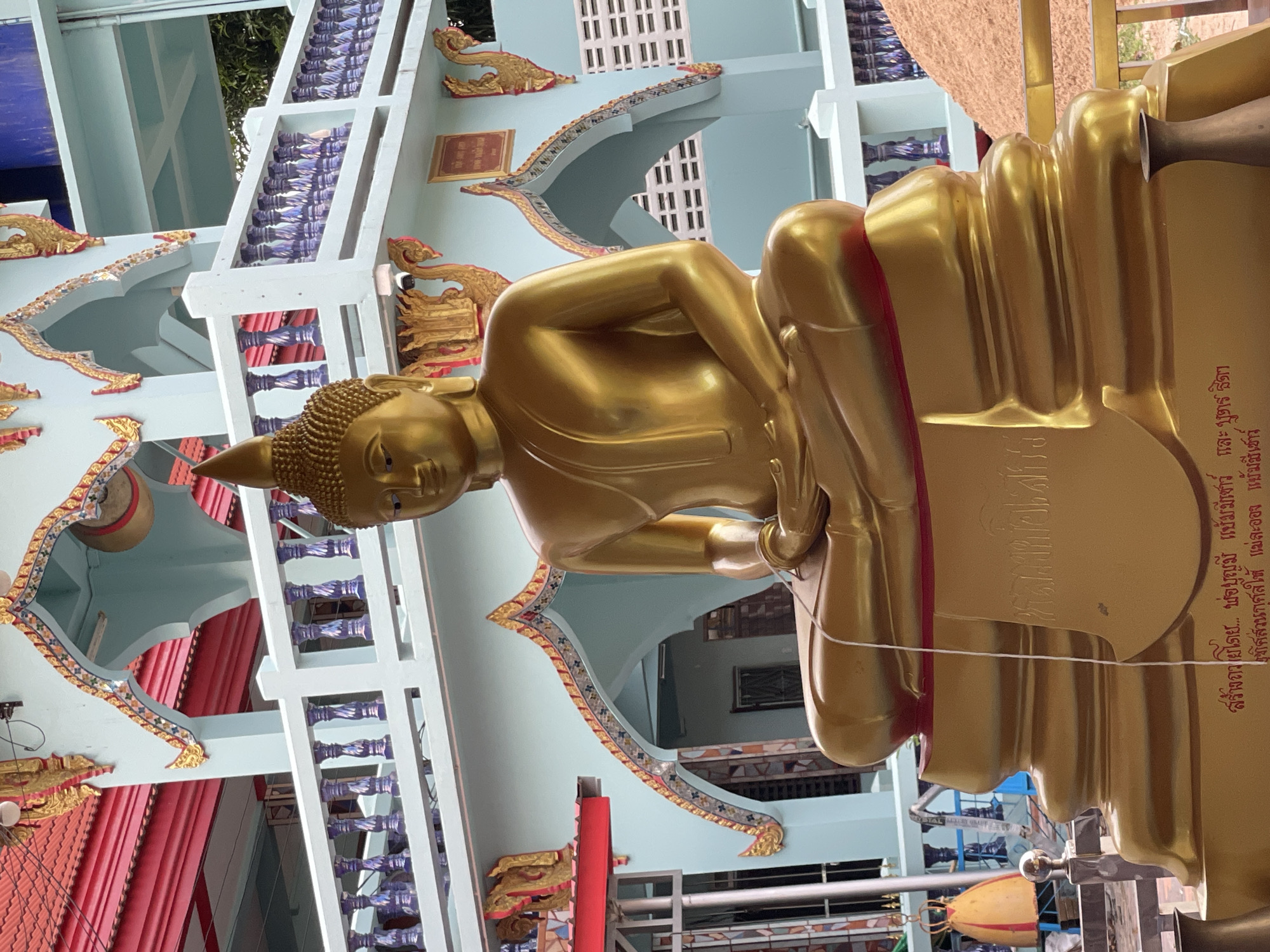 A golden Buddha statue is prominently displayed in the foreground of the image, seated in a meditative pose on an ornate, multi-tiered golden pedestal. The statue faces slightly to the viewer's left, with its eyes closed. A white string drapes from the statue's chest onto the pedestal.
Behind the statue, a light blue building with traditional Thai architectural elements, including decorative golden trim and curved roofs, serves as the backdrop. A white railing with decorative purple balusters runs across the middle section of the building. To the far right of the image, a large pile of sand or dirt is visible.
The scene appears to be outdoors, possibly at a temple or shrine. The statue and the building suggest a location in Pattaya City, Thailand, given the cultural and architectural style. The image is captured from a medium distance, with the statue being the primary subject. There are no people visible in the frame. The lighting suggests it is daytime.