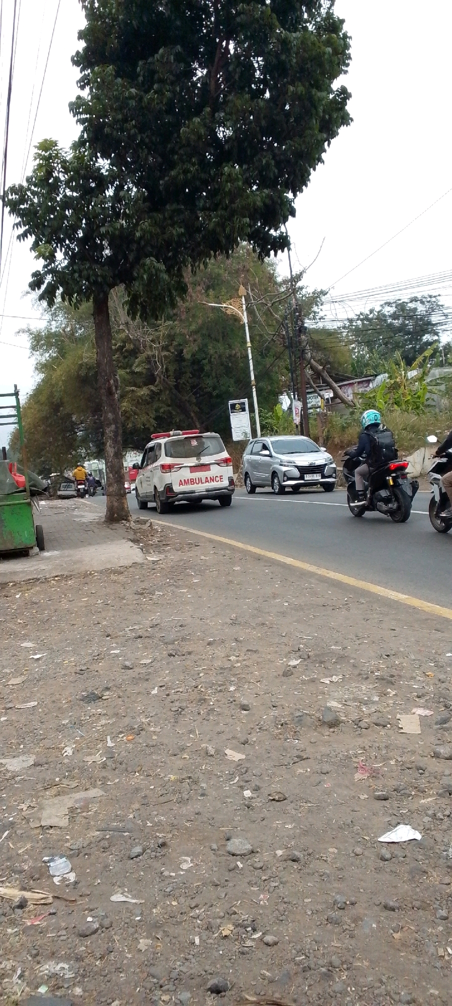 A white ambulance with red flashing lights is parked on the side of a road in Cilaku, Indonesia. The ambulance has "AMBULANCE" written on the back in red lettering. A silver car is driving behind the ambulance, and a motorcycle with a person wearing a blue helmet is in the right lane, also driving. The road is paved and has a yellow line dividing the lanes. The scene appears to be in an urban or suburban area with trees and vegetation on the side of the road. The ground is covered with dirt and debris. The sky is overcast.