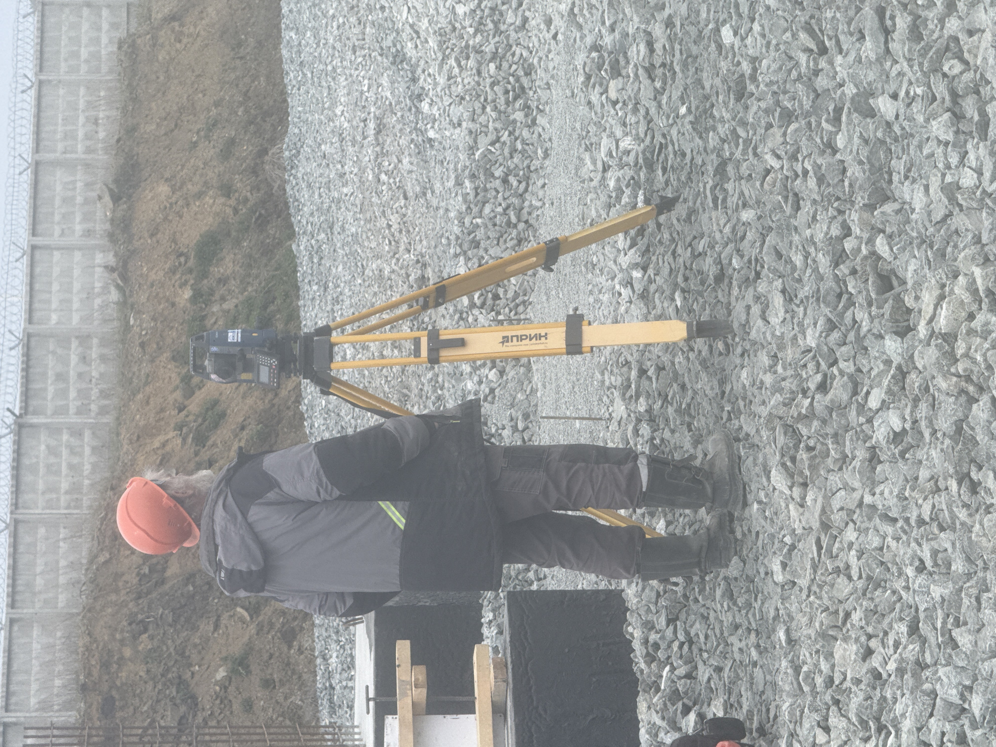 An outdoor scene shows a person wearing an orange hard hat, dark gray work jacket with reflective stripes, dark pants, and black rubber boots standing near a yellow surveyor's tripod with an attached surveying instrument, on a gravel-covered surface. To the person's left, there is a segment of a concrete formwork structure, appearing dark in color. To the left of the person and concrete structure is a dirt slope and a concrete fence topped with barbed wire. The text on the tripod reads "ЯПРИН." This image was taken in the city of Leninsk, Russia.