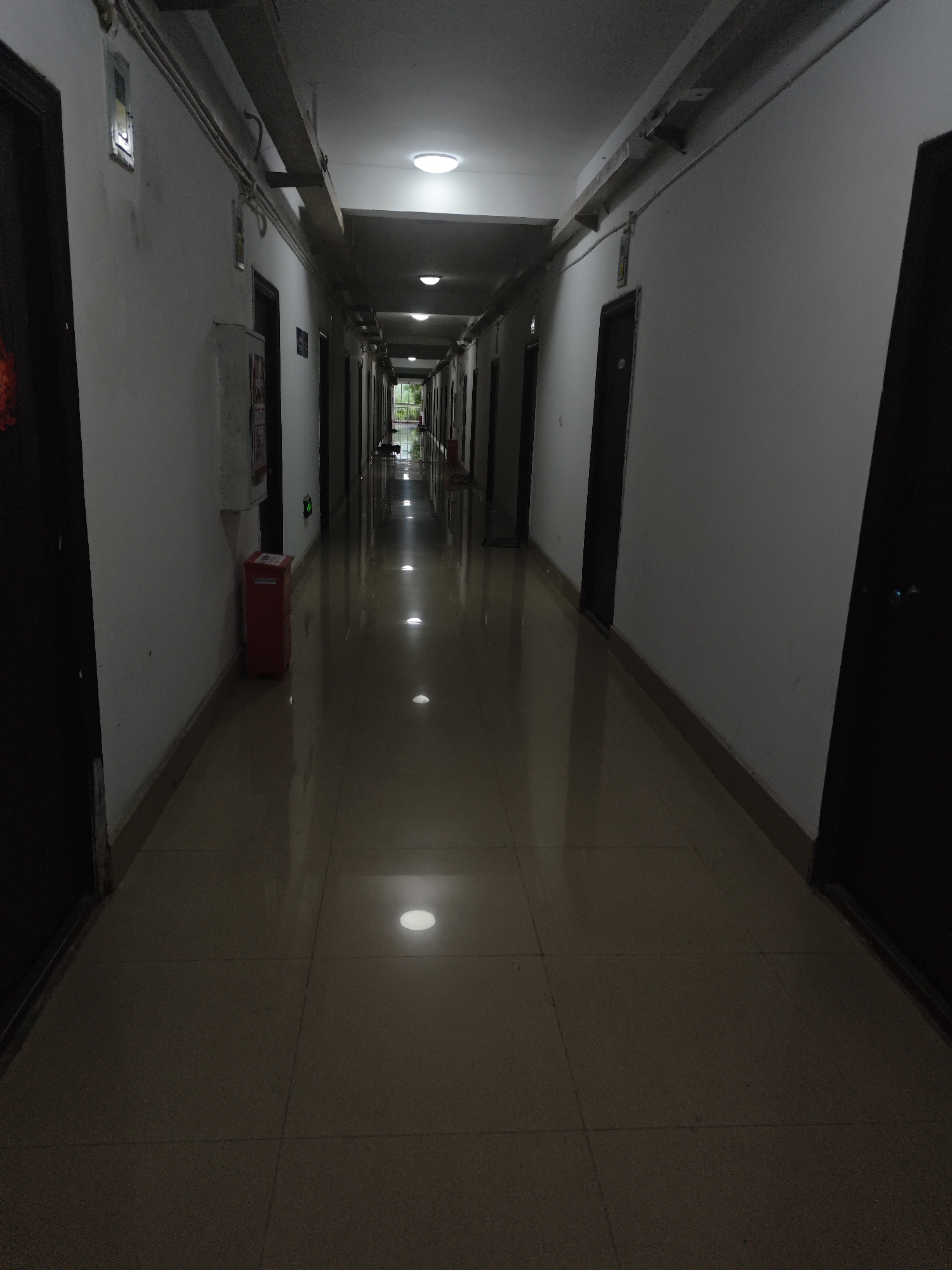 The image shows a long, dimly lit hallway with doors on either side. The hallway has a polished tiled floor that reflects the light from overhead fixtures and recessed floor lights. On the left wall, there is a red fire extinguisher box. The walls are painted white and appear to be slightly textured. The doors are dark, likely wood or metal. The hallway recedes into the distance, where it appears to open up to an outdoor area with greenery visible. The overall impression is of a residential or institutional building hallway, possibly a dormitory or hotel.