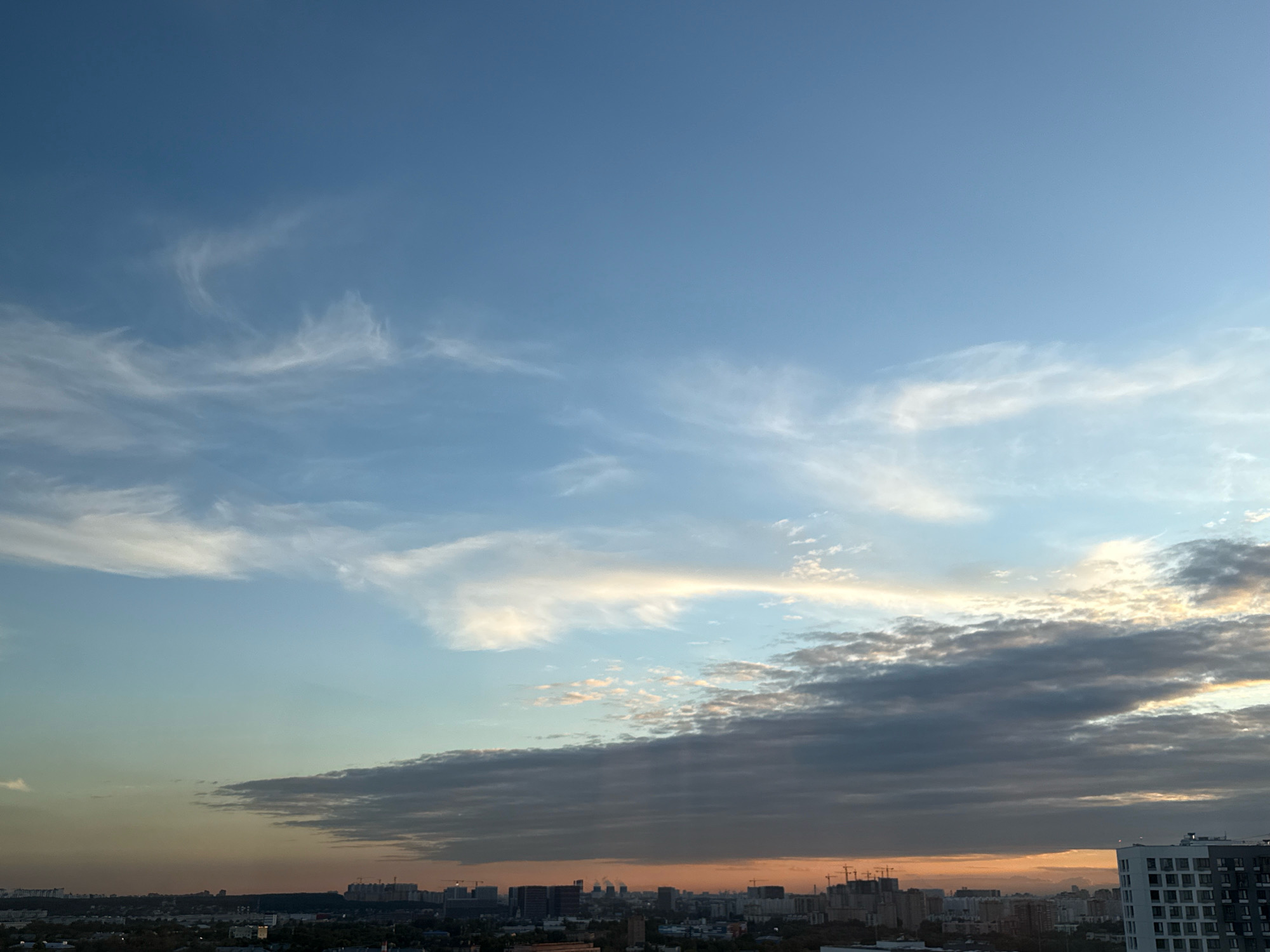 This is a wide-angle, high-angle shot of the sky above Lyubertsy, Russia, during what appears to be either sunrise or sunset. The sky transitions from a clear, bright blue at the top to a warmer, orange-hued gradient closer to the horizon, indicating the presence of the sun below the frame. Wispy, white cirrus clouds are scattered across the upper portion of the sky, becoming more dense and layered towards the horizon, where they are tinged with the orange and yellow light. A band of darker, heavy clouds runs across the middle of the image, casting shadows. Below the clouds, a cityscape is visible, characterized by numerous buildings of varying heights, including apartment blocks and construction sites with cranes. The overall scene is serene and captures the atmospheric conditions of a Russian urban environment at either dawn or dusk. There are no people or animals visible in the frame.