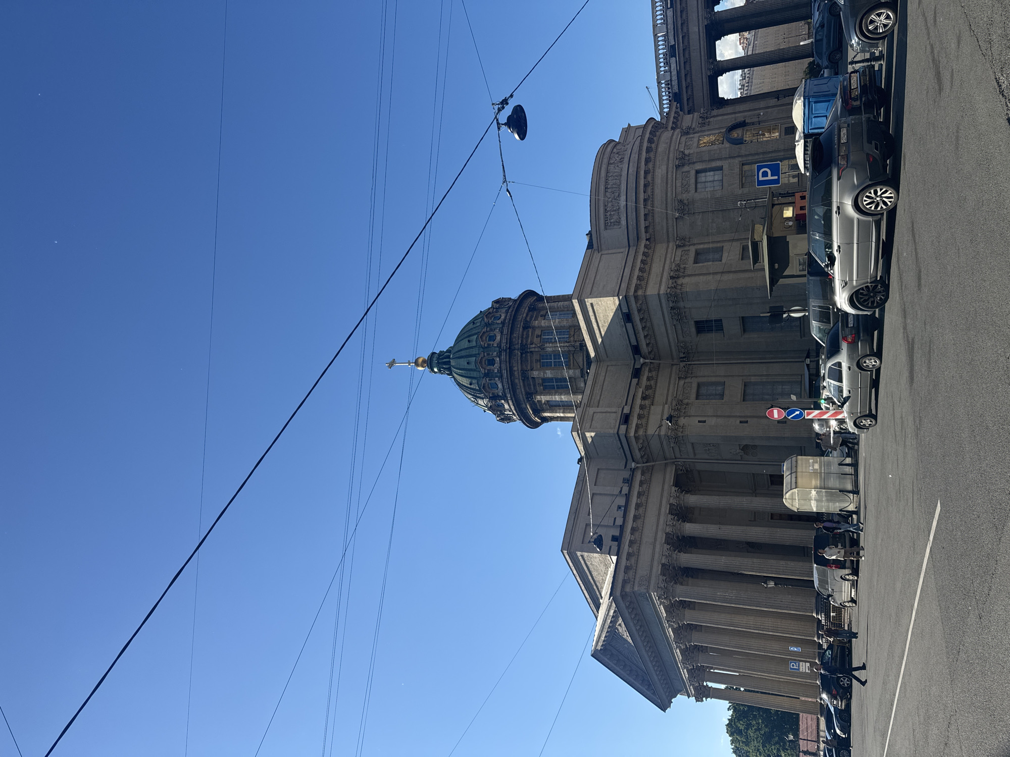 The image shows the Kazan Cathedral in Saint Petersburg, Russia, under a clear blue sky. The majestic cathedral, with its iconic green dome and classical colonnade, dominates the scene. Cars are parked along the street in front of the building, and a few people can be seen walking on the sidewalk. The time of day appears to be midday, judging by the bright sunlight casting shadows. The weather is sunny and clear. Overhead, a network of wires crisscrosses the sky, a common sight in urban environments. The overall atmosphere is one of peaceful urban life, with the grandeur of the historical architecture providing a striking backdrop. Visible text includes a blue parking sign with a white "P", and a traffic sign with a red circle and a horizontal blue line.