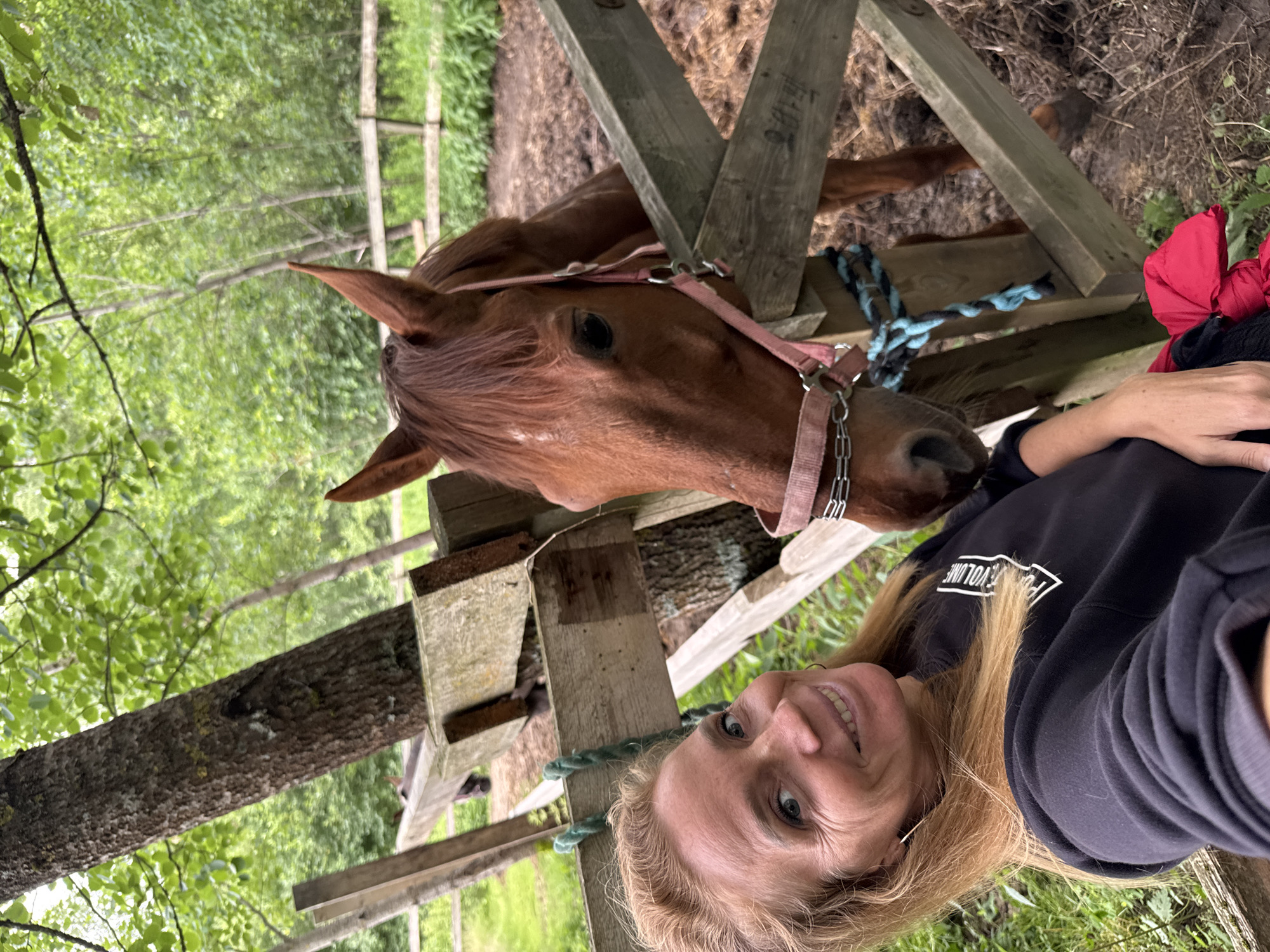 An outdoor selfie depicts a smiling blonde woman in a black sweatshirt standing next to a brown horse. The horse, wearing a pink halter and a blue and black lead rope, is partially visible behind a wooden fence. The woman has blonde hair and is wearing hoop earrings. The background shows trees and a wooden fence. The scene appears to be a stable or farmyard, though no specific location in Mars, Russia is identifiable.