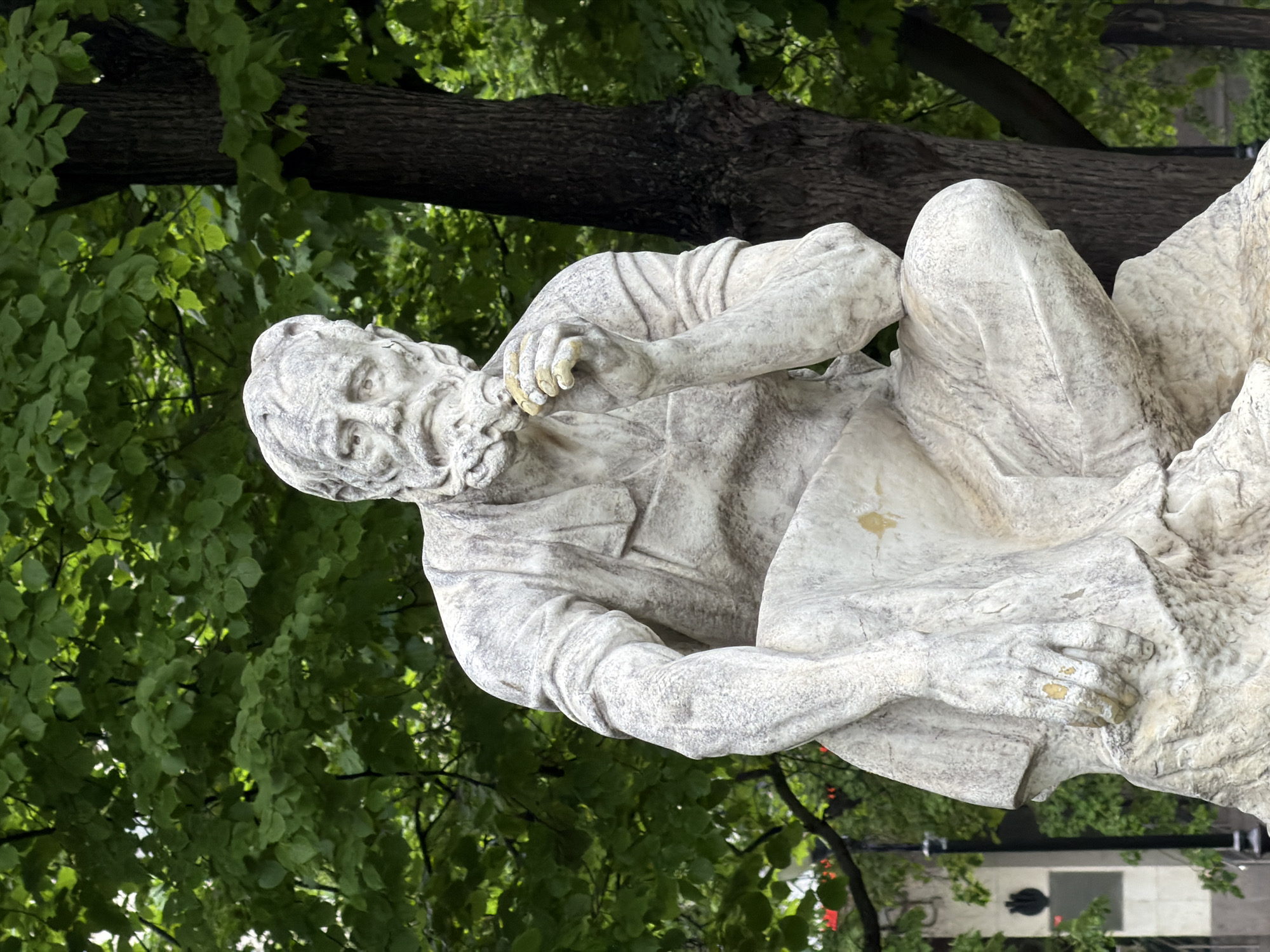The image shows a statue of a seated man with a beard, likely a historical or literary figure. The statue is made of a light-colored stone or concrete. It is situated outdoors, with a background of green foliage and tree trunks. In the background, glimpses of a street and buildings can be seen, suggesting an urban park or square location. Based on the style of the statue and the general environment, it is likely located in the city of Moscow, Russia.