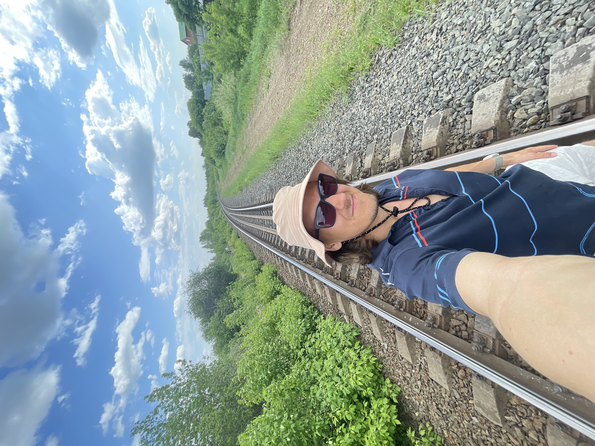A man lies on a railway track. The railway is located in a rural area near Izhevsk, Russia, as indicated by the landscape. The man is wearing a hat, sunglasses, and a dark-colored shirt with blue stripes. He is positioned in the middle of the tracks, holding his arm out as if taking a selfie. The sky is blue with white clouds. Green vegetation lines both sides of the railway. The tracks themselves are made of steel rails supported by concrete ties. Gravel fills the space between the ties.