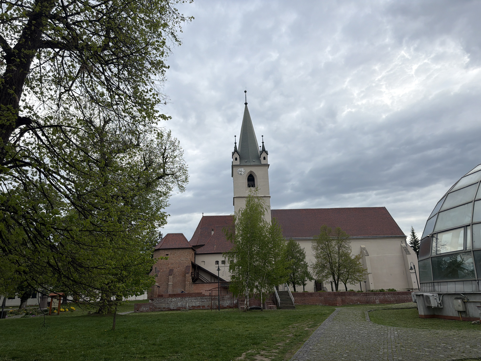 An exterior long shot captures a fortified church complex under a dramatic, overcast sky. The central subject is the church, featuring a tall, light-colored bell tower topped with a pointed spire and four smaller turrets at its base. A clock face is visible on the tower, though the exact time is not discernible. The church building itself has light-colored walls and a distinct red-tiled roof, with various attached structures made of reddish brick, suggesting a historical, possibly fortified, architectural style.
In the immediate foreground on the left, a large, deciduous tree with fresh, light green leaves partially frames the scene, its branches extending towards the top and center of the frame. The ground beneath is covered in lush green grass, with a winding cobblestone path visible on the right side, leading towards the church. Other smaller trees are scattered around the complex, also showing new spring foliage.
To the far right, contrasting sharply with the historic architecture, is a modern, spherical or dome-shaped glass structure with visible windowpanes and what appear to be air conditioning units at its base. The sky above is entirely covered with thick, textured gray clouds, indicating an overcast day. No human activity is explicitly visible, lending a quiet, contemplative atmosphere to the scene. The overall setting is an outdoor space, possibly a park or courtyard surrounding the historical church. The architecture hints at a location within Transylvania, aligning with Târgu Mureș, Romania.