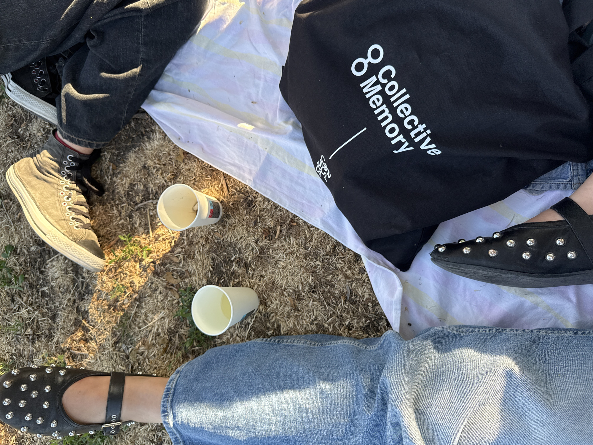 Two individuals are seated outdoors on a patch of dry grass, possibly a park, with a picnic blanket spread between them. One individual on the left wears distressed light-wash denim jeans and a black slip-on shoe adorned with silver studs and a buckle. Their right leg is crossed over their left. Between them, on the ground, are two disposable white cups, one appearing to contain a light-colored liquid and the other a crumpled white object. To the right, the other individual is dressed in dark wash jeans and grey high-top sneakers with white laces. A black tote bag with white text reading "Collective Memory" is positioned on the picnic blanket. The sun casts dappled shadows on the ground, suggesting it is daytime and outdoors. The location is not identifiable as Sofia, Bulgaria from the visual information provided.