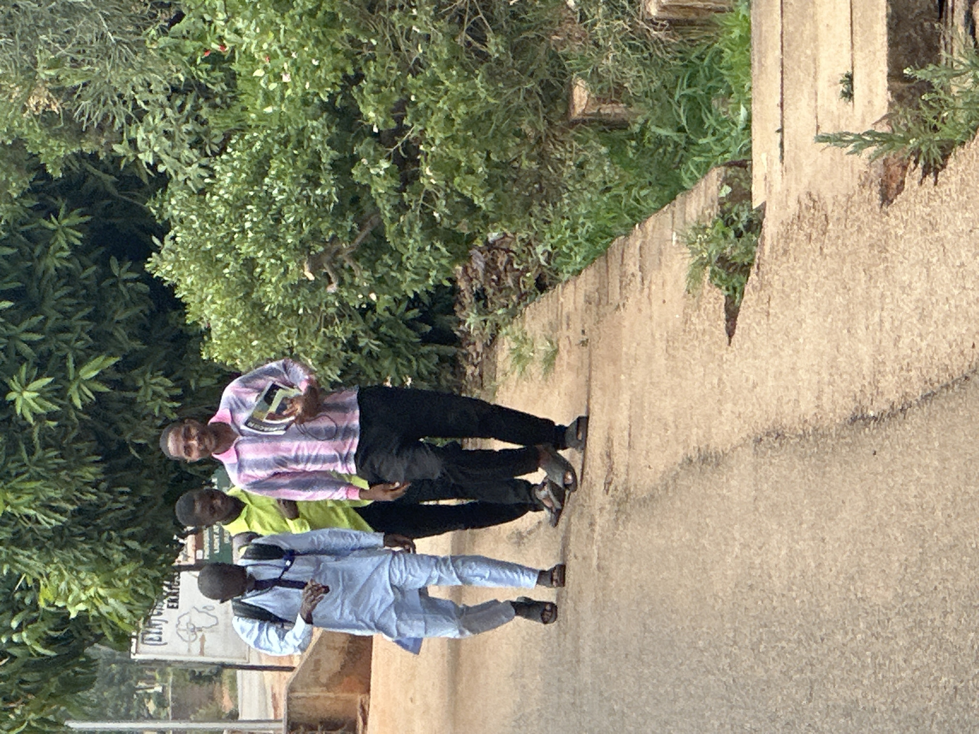 Three males stand near a walkway beside dense foliage in Bukuru, Nigeria. The man closest to the camera wears a light blue outfit with a black backpack and a black tie. The next man wears a bright green shirt. The third man holds a book in his left hand and wears a pink and white striped shirt with black pants. All three wear sandals. A sign that reads "CENT LIGHT A EXAM" is partially visible. The walkway is made of concrete and has plants growing out of its edge.