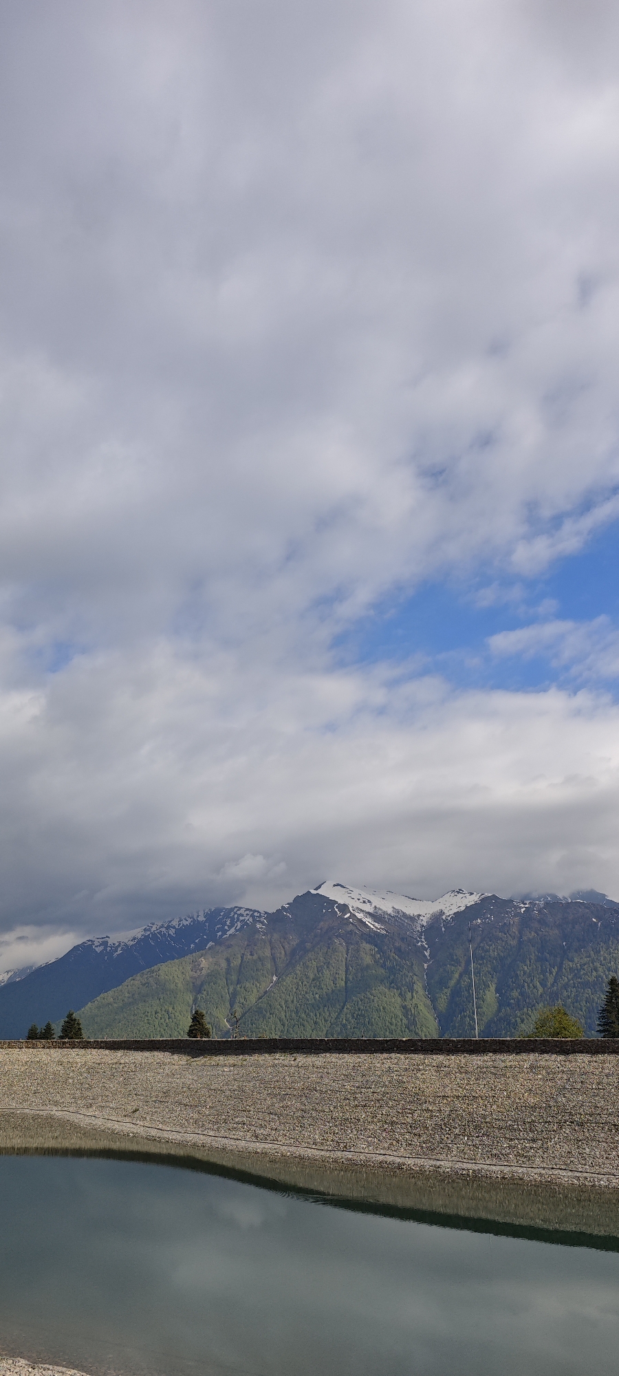 The image shows a mountain range with snow-capped peaks partially obscured by clouds under a sky with mixed clouds and blue patches. In the foreground, there's a structure, possibly a dam or reservoir, made of stonework, with a body of water reflecting the sky and parts of the structure. Several green trees stand near the crest of the structure. The location is Unknown, Russia.