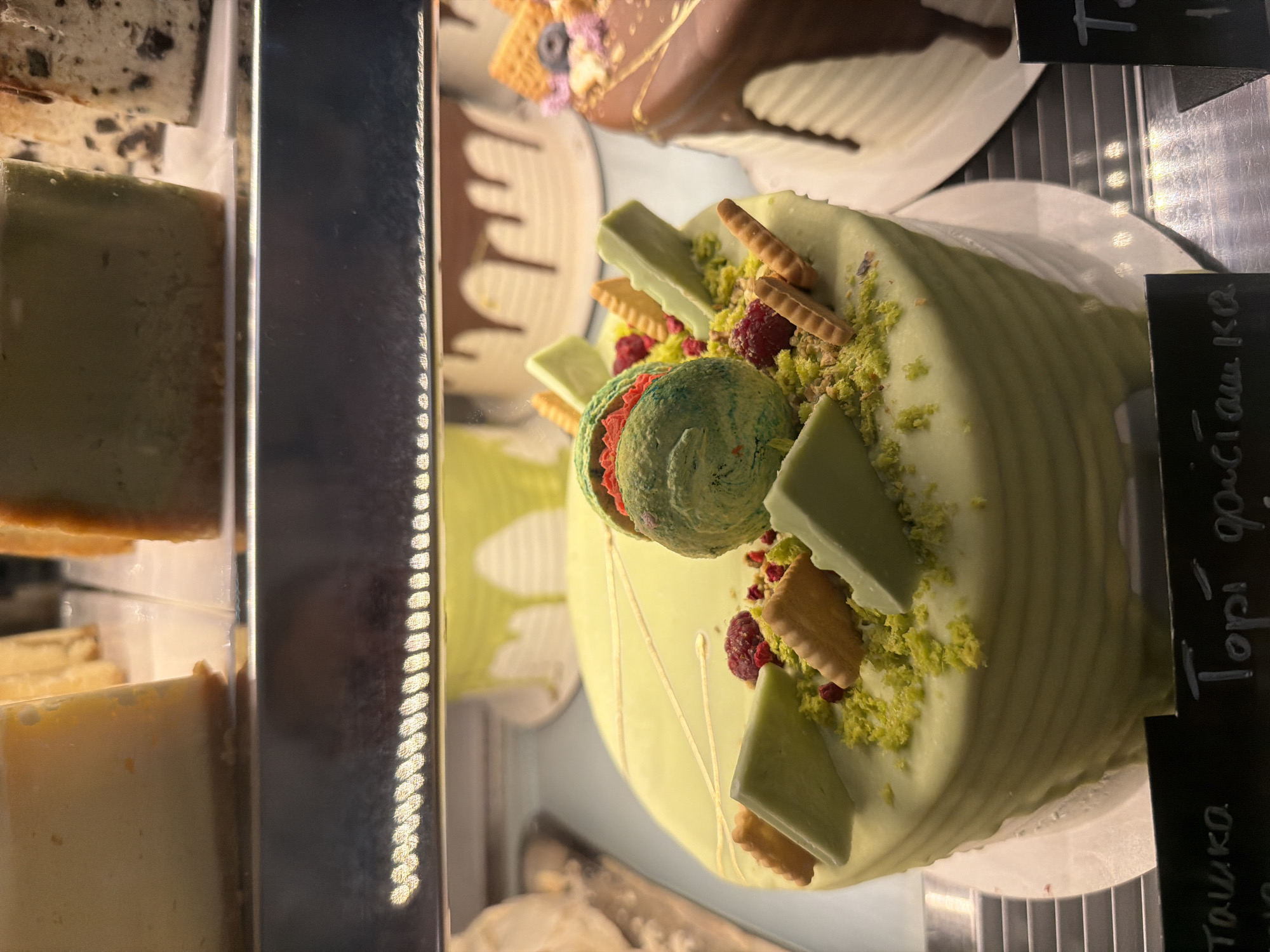 The image shows a close-up of a light green cake with various decorations. The cake is round and has a rippled texture on its sides, suggesting it might be a mousse or layered cake. It is topped with pieces of green chocolate or frosting, small crackers, and red berries, possibly raspberries. There is also a decorative swirl of green and red frosting or ice cream in the center.
The cake is displayed in what appears to be a refrigerated display case, as indicated by the reflective surfaces and the vertical metal grating visible in the background. Other desserts or food items are partially visible in the background and to the left, including what looks like slices of cake or pastries.
Two black labels with white writing are visible on the right side of the cake. One label, partially obscured, appears to say "Tацшка." The other, more visible label, says "Tорi дочіашка." The writing is in a stylized font. The overall lighting suggests an indoor setting, likely a bakery or dessert shop. The image is taken from a slightly elevated angle, focusing on the decorated cake. There are no people visible in the image.