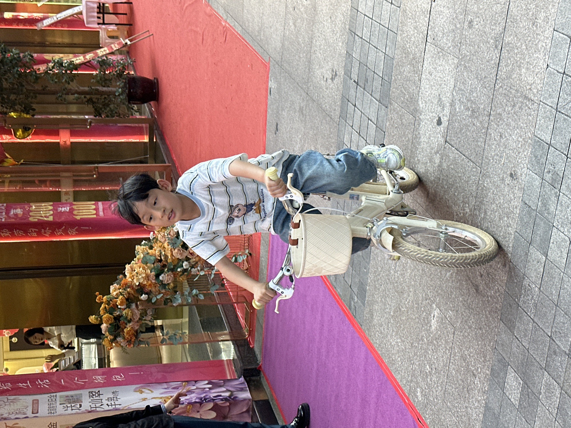 A young boy in a striped t-shirt and jeans is riding a cream-colored bicycle with a basket in the street. He is holding the handlebars and looking at the camera. The scene is in Jiangjin, China, evidenced by the Chinese writing on the building signage behind him. The boy is riding on a tiled sidewalk adjacent to a building with a red carpet, a purple carpet, and flower decorations. A person in a black suit stands near the purple carpet. There is also a person inside the building that is visible through the glass.
