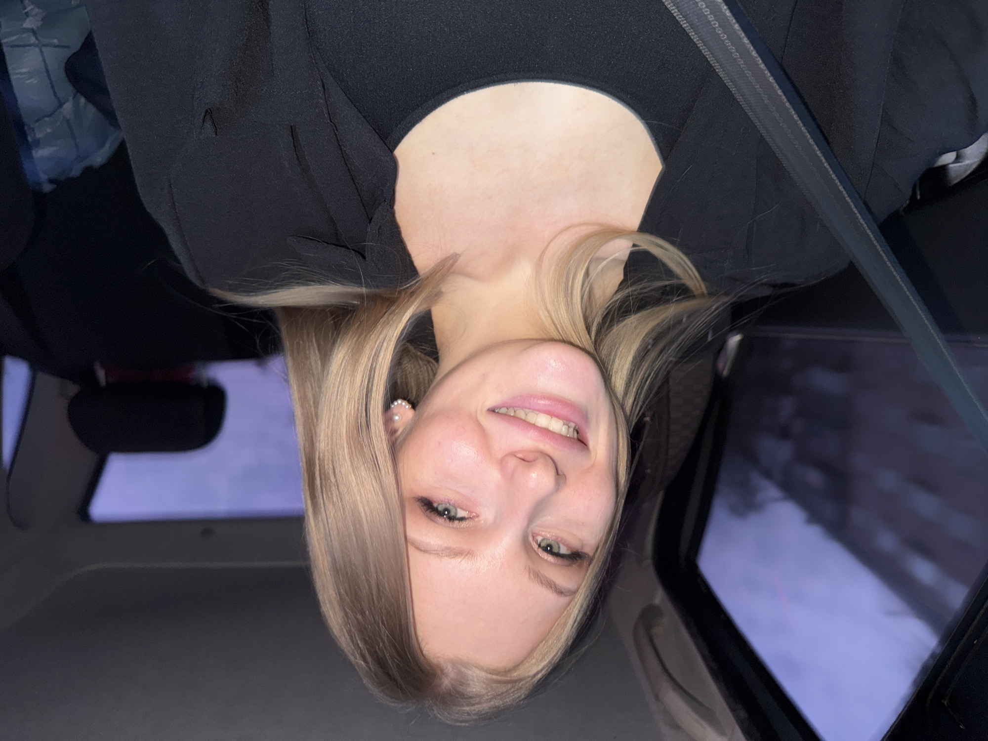 A young woman with blonde hair is photographed from a low angle inside a car. She wears a black top, and the seatbelt is visible diagonally across her torso. Outside the car windows, a blurred cityscape suggests it may be nighttime or dusk. The image was taken in Perm, Russia, based on the prompt.