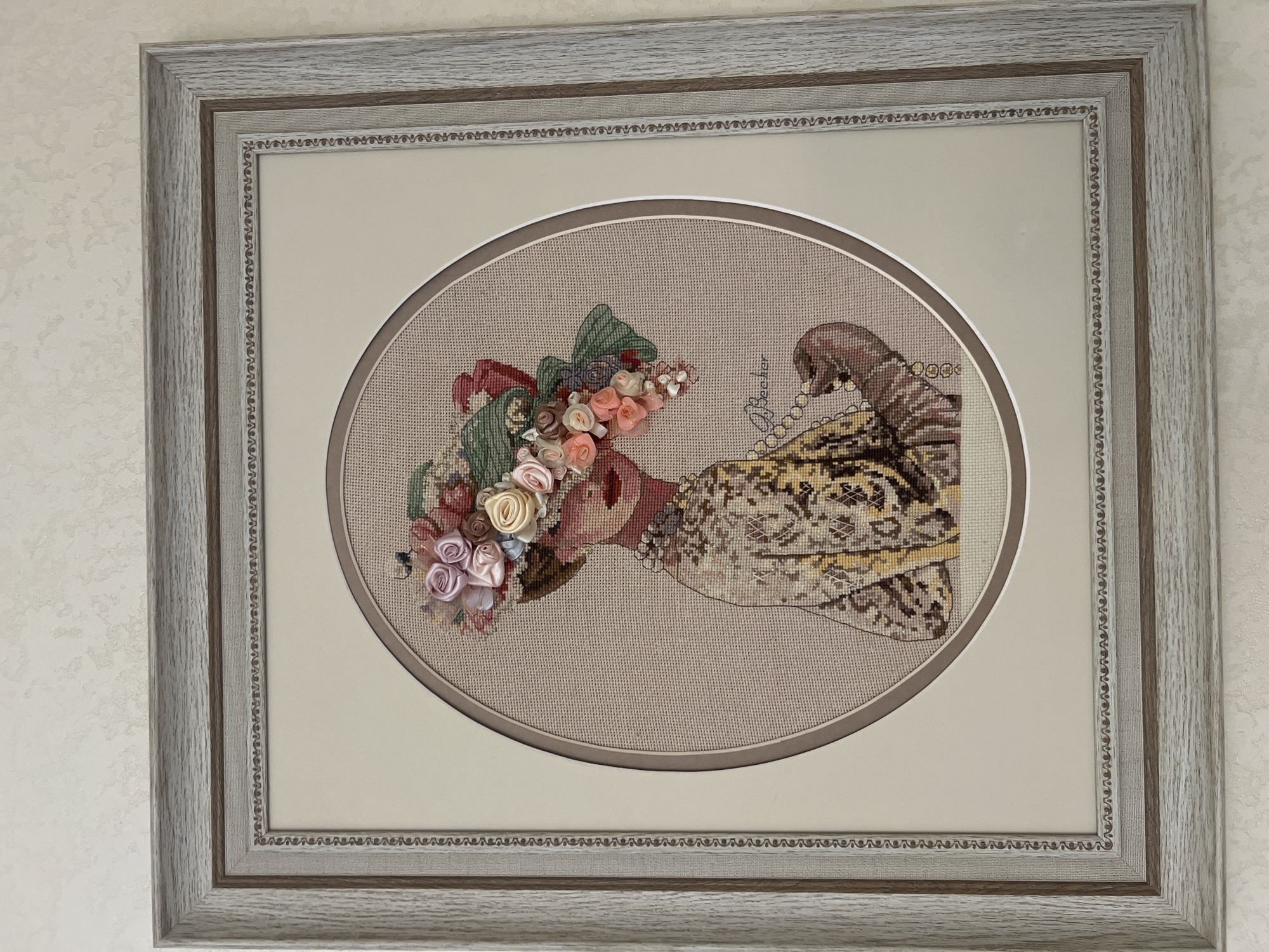 A framed oval cross-stitch artwork depicts a woman from the shoulders up, wearing a wide-brimmed hat adorned with various ribbon roses and green ribbon leaves. The woman has fair skin, red lips, and appears to be wearing a pearl necklace. Her dress has a patterned design in shades of yellow, brown, and white. She is holding a string of pearls in her right hand. The artwork is signed "J. Becker" in cursive handwriting. The cross-stitch is mounted on a light beige mat board, which is then placed within a light gray wooden frame with a textured surface. The frame has a narrow, decorative inner border with a repeating pattern. The artwork is displayed on a wall with textured off-white wallpaper. The image appears to have been taken indoors, possibly in a residential setting in Bender, Moldova.