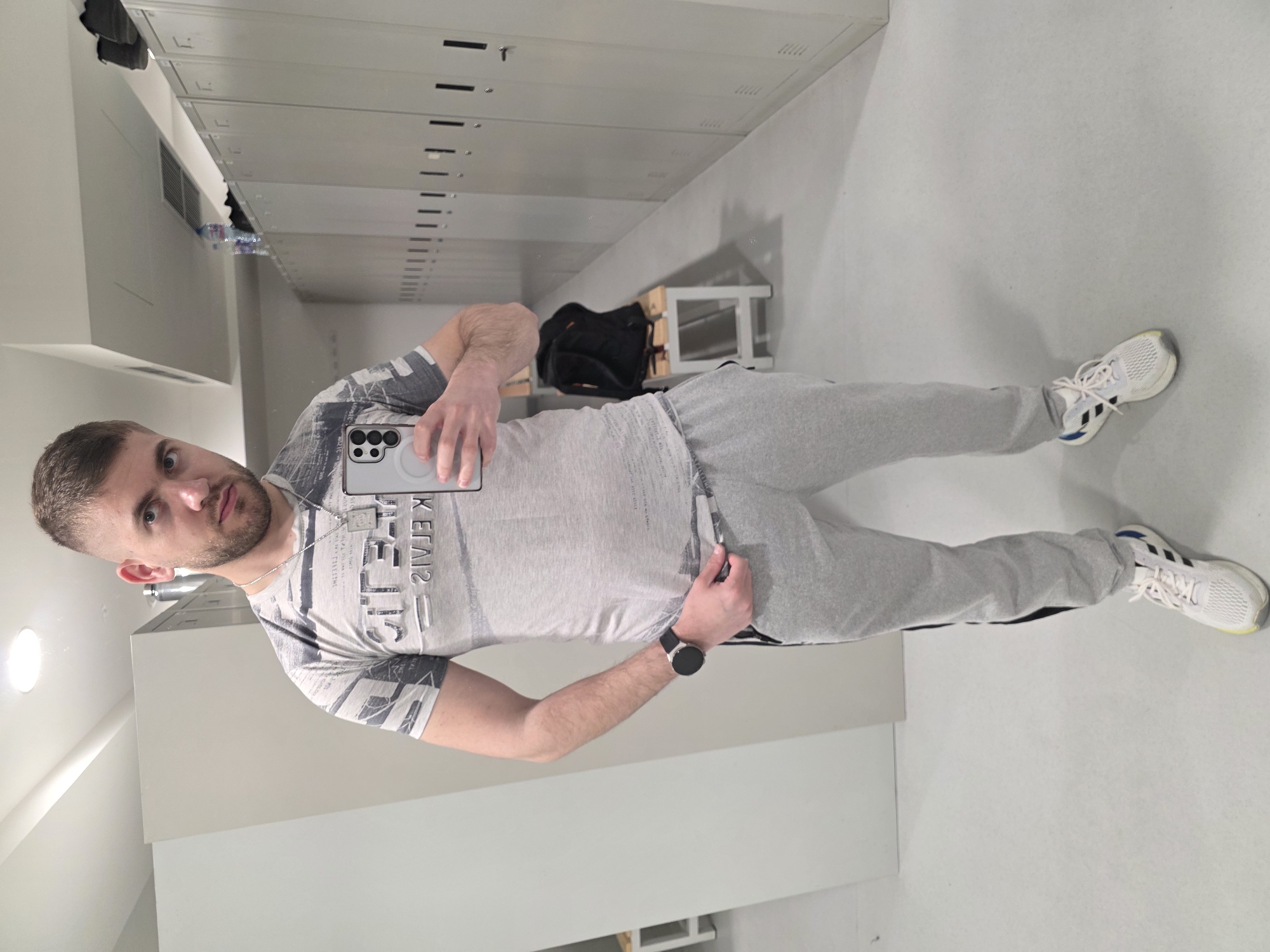A man is taking a selfie in what appears to be a locker room. He is wearing a grey t-shirt, grey sweatpants, and white sneakers with black stripes. He is holding a smartphone with a prominent camera array. He is wearing a black smartwatch on his left wrist. Behind him are rows of lockers, a bench, and a black bag. The location is unspecified and not verifiably Plovdiv, Bulgaria based solely on the provided visual information.