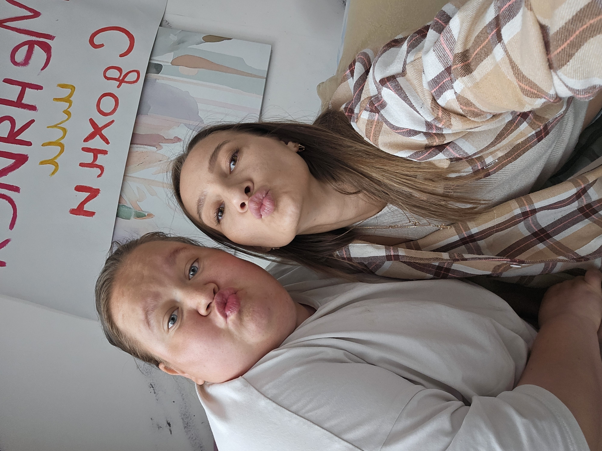 This image depicts two adult females posing for a selfie indoors, located in what is stated as the city of Chaikovsky, Russia. Both individuals are looking directly at the camera with their lips pursed into a "kiss" pose. The subject on the left has light skin, blue eyes, and light brown hair pulled back. She is wearing a plain light-colored, possibly white or light grey, short-sleeved t-shirt. The subject on the right has light skin, brown eyes, and long brown hair. She is wearing a light-colored, ribbed long-sleeved top underneath a plaid button-up shirt featuring shades of beige, brown, white, and light pink. She also wears a delicate necklace with a small pendant and small hoop earrings. The two subjects are positioned closely together, suggesting a friendly or intimate interaction.
The background consists of a light-colored wall. On the left side of the frame, the wall is mostly plain, showing some dark smudges or markings in the upper left corner. On the right side, there is a white poster or sheet of paper affixed to the wall, displaying handwritten text in red and yellow. The red text, which appears to be in Cyrillic, includes characters such as "Н", "Р", "Х", "В", "О", "С", and "Я", and is oriented in a manner that appears reversed or upside down. Yellow wavy lines are drawn beneath some of the red letters. Below this poster, another piece of artwork or paper with abstract pastel shapes in hues of blue, green, beige, and pink is visible. The right subject appears to be seated on or leaning against a light-colored, possibly beige or tan, upholstered piece of furniture, such as a couch or chair.