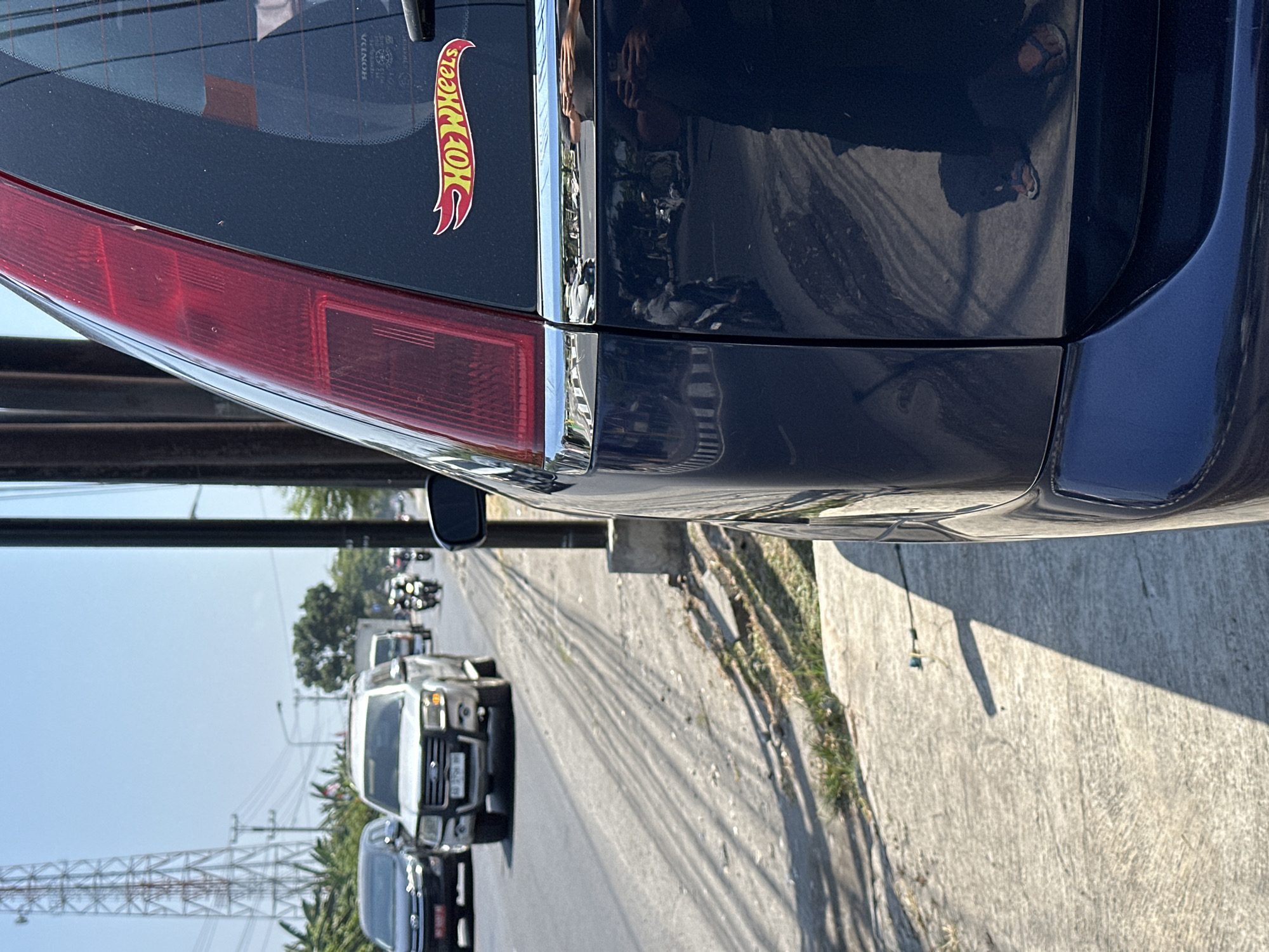The image shows a dark blue car parked on the side of a street in Bulakrejo, Indonesia. The car is viewed from a low angle, with its rear right side dominating the frame. A red taillight is visible on the top left of the car. On the rear windshield, a "Hot Wheels" logo sticker is placed on the right side. The car's paint reflects the surrounding environment, showing a street with traffic, including other cars and motorcycles. A sidewalk runs alongside the street, with some dry grass growing through cracks. In the background, a tall transmission tower can be seen, along with trees and buildings, indicating an urban or suburban setting. The sky is clear and blue, suggesting a sunny day.