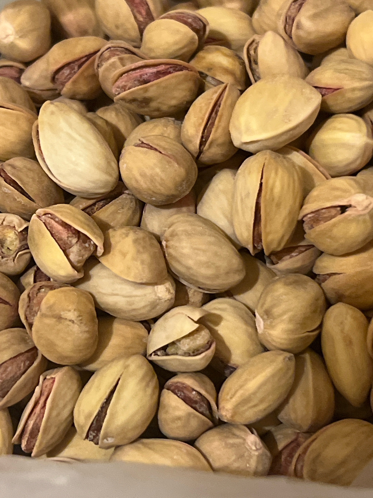 A close-up, top-down shot captures a pile of unshelled pistachios. The pistachios are a light tan color with some exhibiting reddish-brown hues visible through their split shells. They are tightly packed together, filling the frame, and appear to be a food item. The lighting is even, and the focus is sharp throughout the image. No specific location or environmental details are discernible.