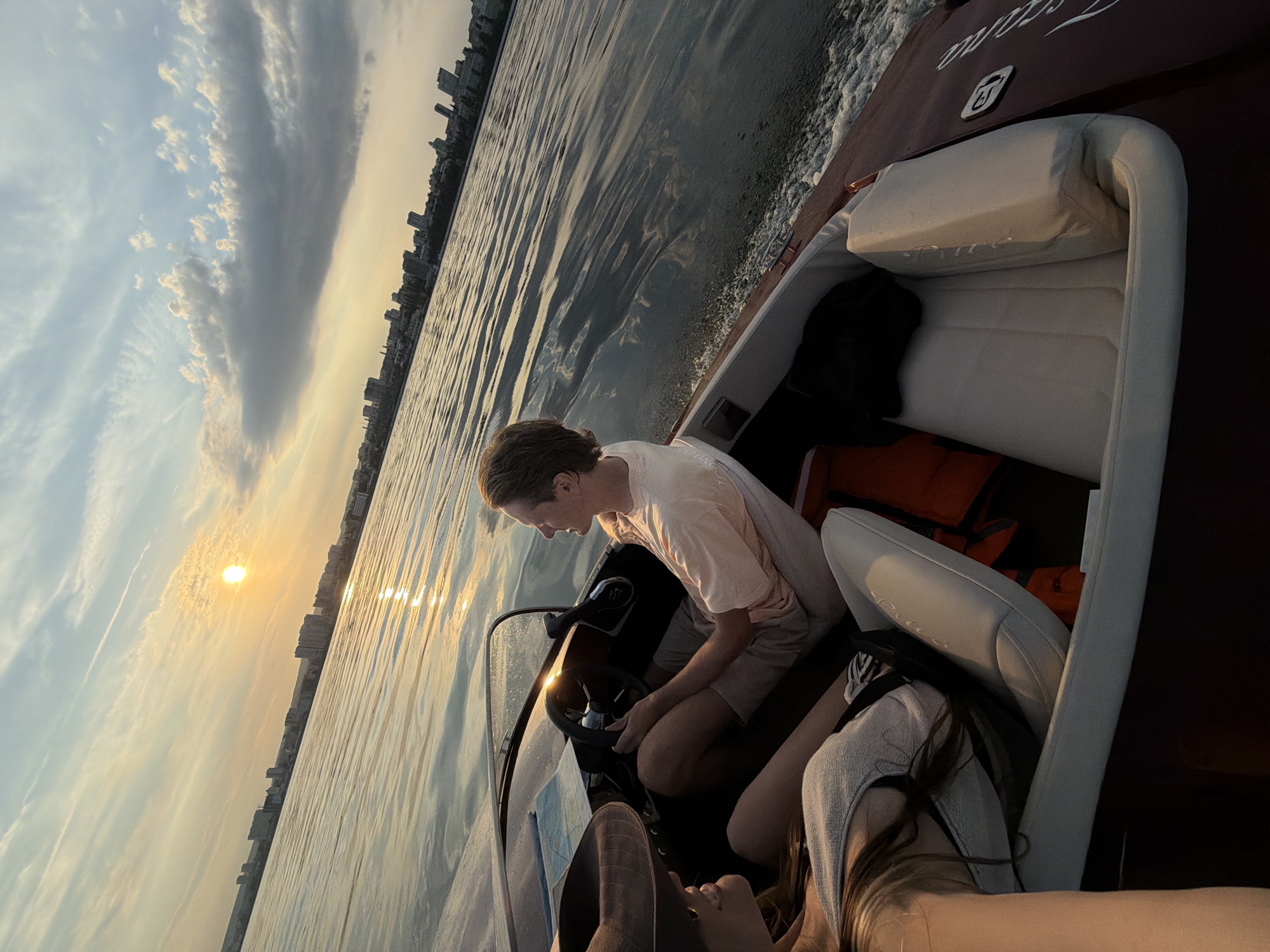 Two individuals are aboard a maroon motorboat cruising on a wide body of water, likely the Volga River, with the Saratov skyline visible in the distance. The sun is setting, casting a warm glow across the sky and reflecting on the rippling water. The person on the left, wearing a wide-brimmed hat and a light-colored top, is partially visible, facing the person on the right. The person on the right, a young male with light brown hair, is steering the boat, wearing a peach-colored t-shirt and light-colored shorts. He is looking to his right, seemingly at the other occupant. The interior of the boat features white upholstered seating. Life vests are visible in the boat, one orange and one black. The boat is moving, creating a wake behind it. The city skyline features numerous buildings of varying heights.