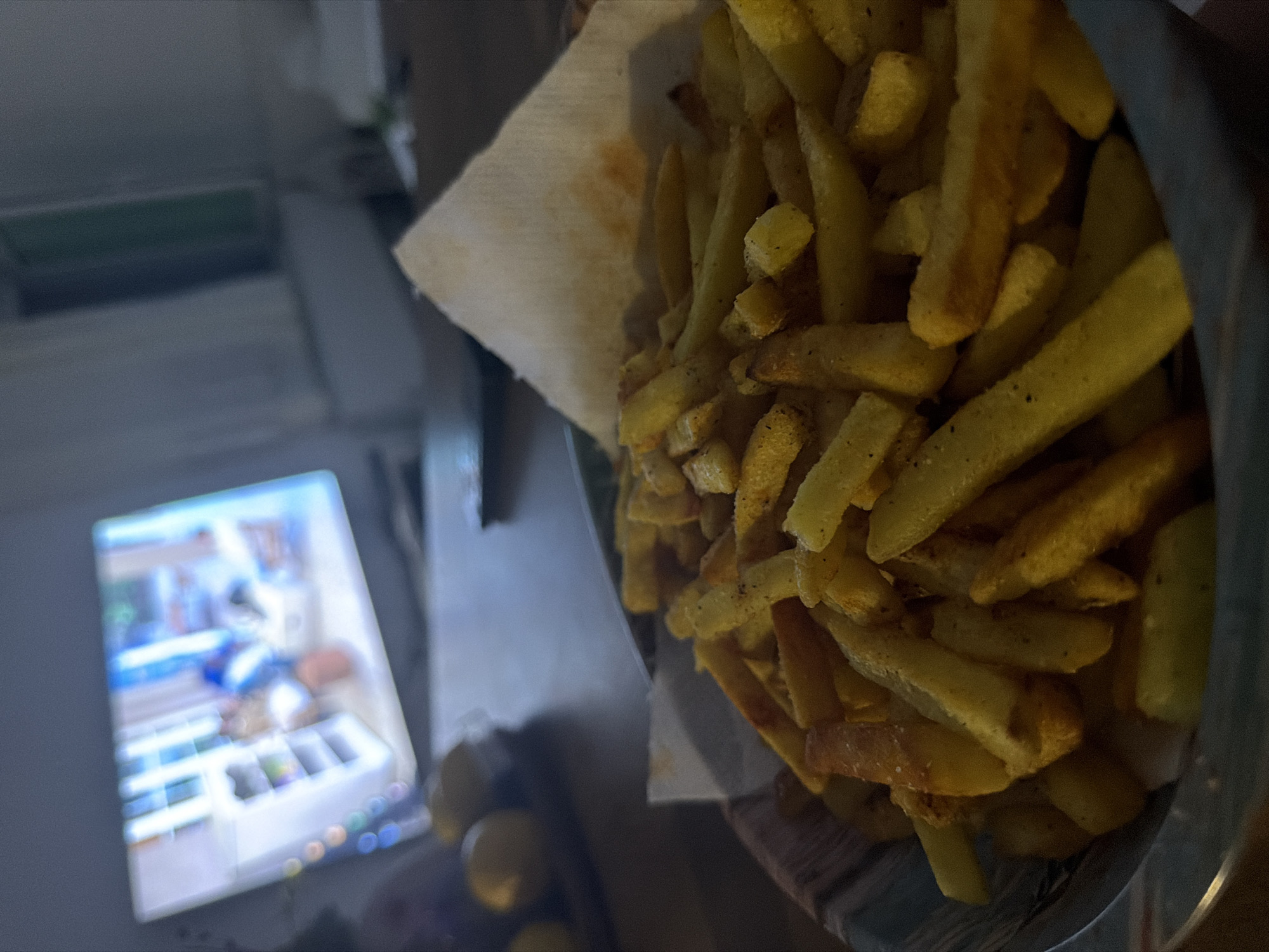 A close-up, angled shot focuses on a bowl of golden-brown french fries, seasoned with salt and pepper. The fries are piled high in a striped bowl, with a white paper napkin peeking out from the top. In the blurred background, a television screen displays a scene of a brightly lit room with shelves and what appears to be furniture. Two yellow, round objects, possibly lemons, are visible in the middle ground on the left side of the image. The overall lighting is dim, suggesting an indoor evening setting. The scene does not contain any information that would allow for its location to be identified as Shiroka Poliana, Bulgaria.