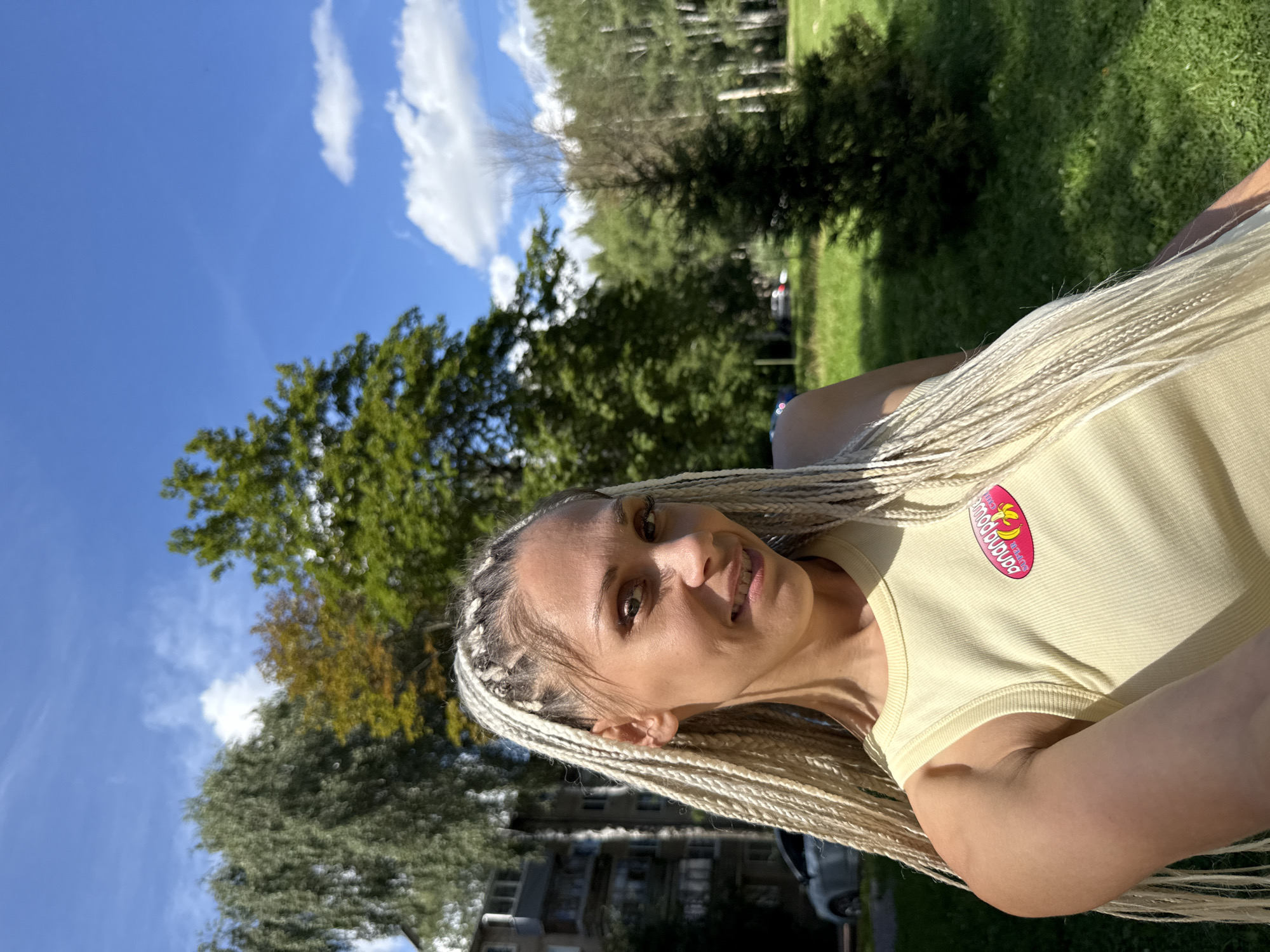 In Rusanovo, Russia, on a sunny day, a woman with blonde braided hair is taking a selfie outdoors. She is wearing a yellow tank top with a pink and yellow oval-shaped logo that reads "banana power." The background consists of green grass, trees, a building, and a blue sky with scattered white clouds.