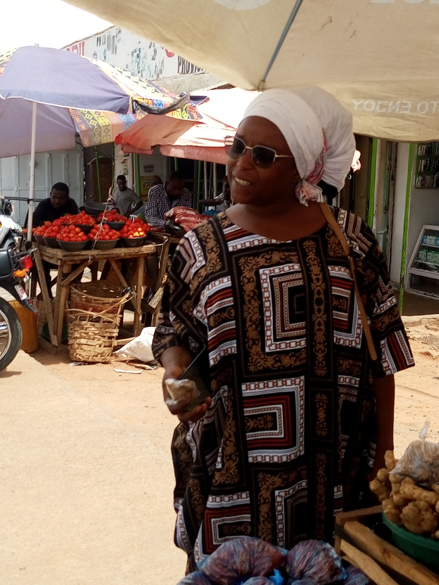 The image depicts a market scene in Tempure, Nigeria. A woman wearing a white headscarf, sunglasses, and a patterned dress stands in the foreground. She holds an object in her hand. Behind her, to the right, are vegetables and what appears to be ginger. In the background, vendors sit behind tables piled high with tomatoes, under colorful umbrellas. Additional people and market stalls can be seen further in the background, and a motorcycle is parked on the left. The scene is outdoors on a sunny day.