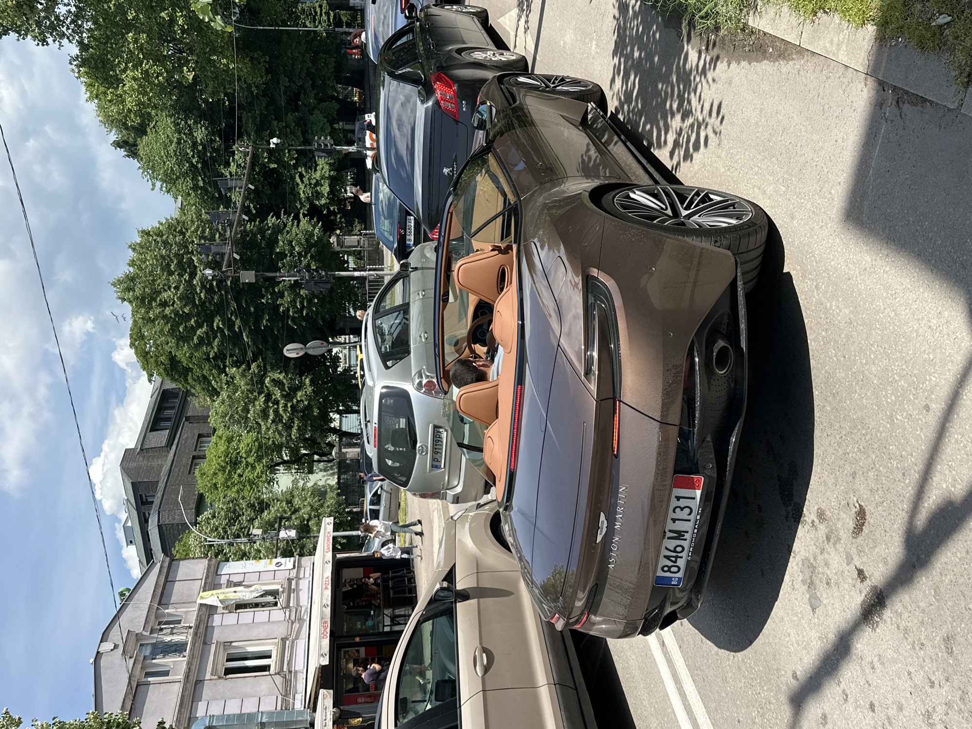 The image shows a street in Varna, Bulgaria, with several parked cars. A brown Aston Martin convertible is the most prominent vehicle, facing away from the viewer. The license plate number is 846 M 131. Other cars include a silver Toyota, a beige car, and a dark grey Peugeot. A building with signs for "Döner," "Pizza," and "Sandwich" is visible. People are walking on the sidewalk. Trees line the street, and a traffic light is visible. The weather appears sunny.