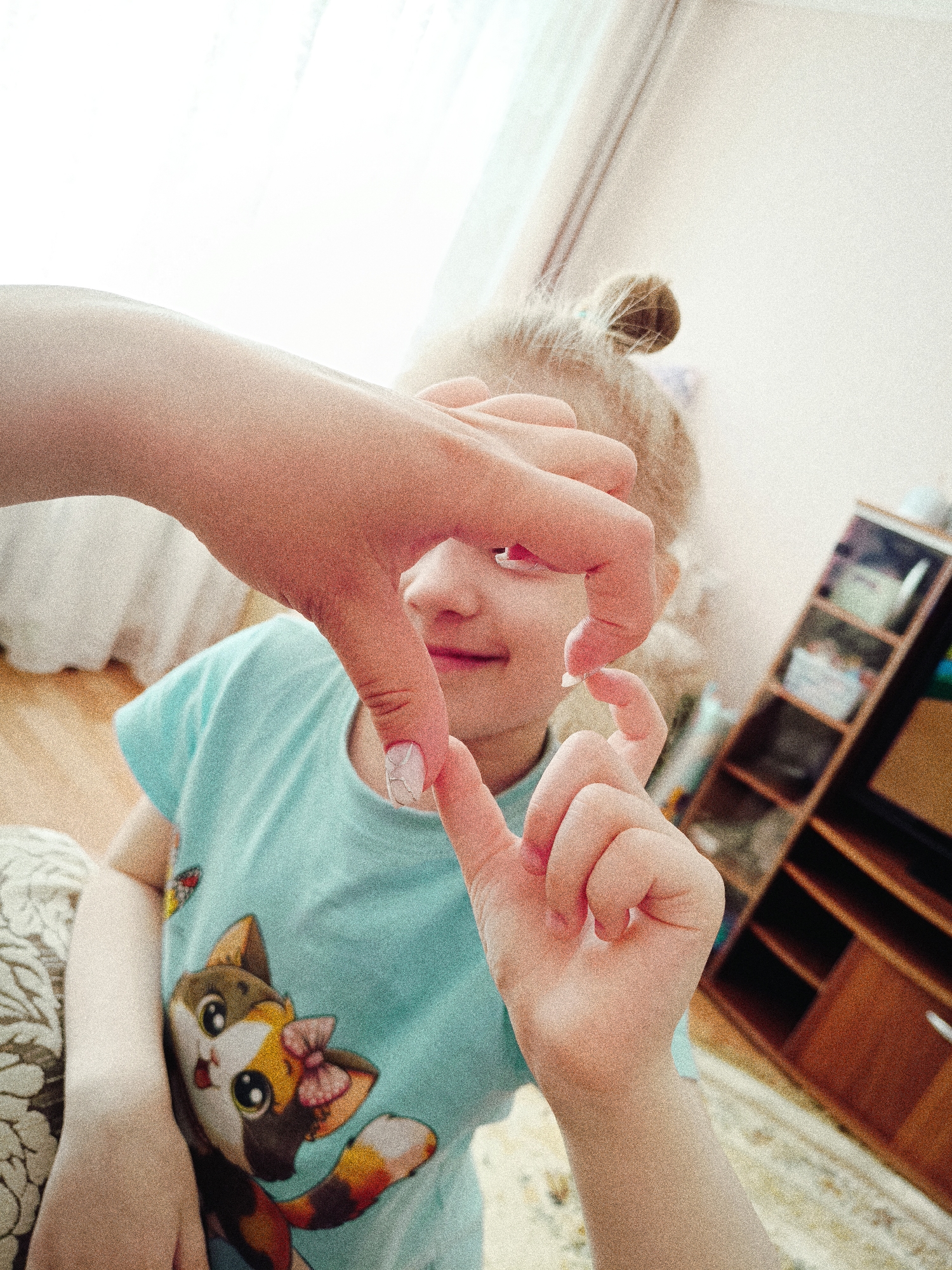 A close-up, high-angle shot depicts a child, appearing to be a young girl with fair skin and blonde hair styled in a bun, sitting indoors with a hand holding up a frame in the shape of a heart in front of her face. The girl is wearing a light blue shirt with a cartoon cat graphic on the front. Behind her is a partially visible wooden shelf unit filled with various items. The background is brightly lit, with a window and curtain visible. The photo appears to have been taken indoors, with a carpet visible in the lower right. Based on the metadata, the location of this image is in Tobolsk, Russia.