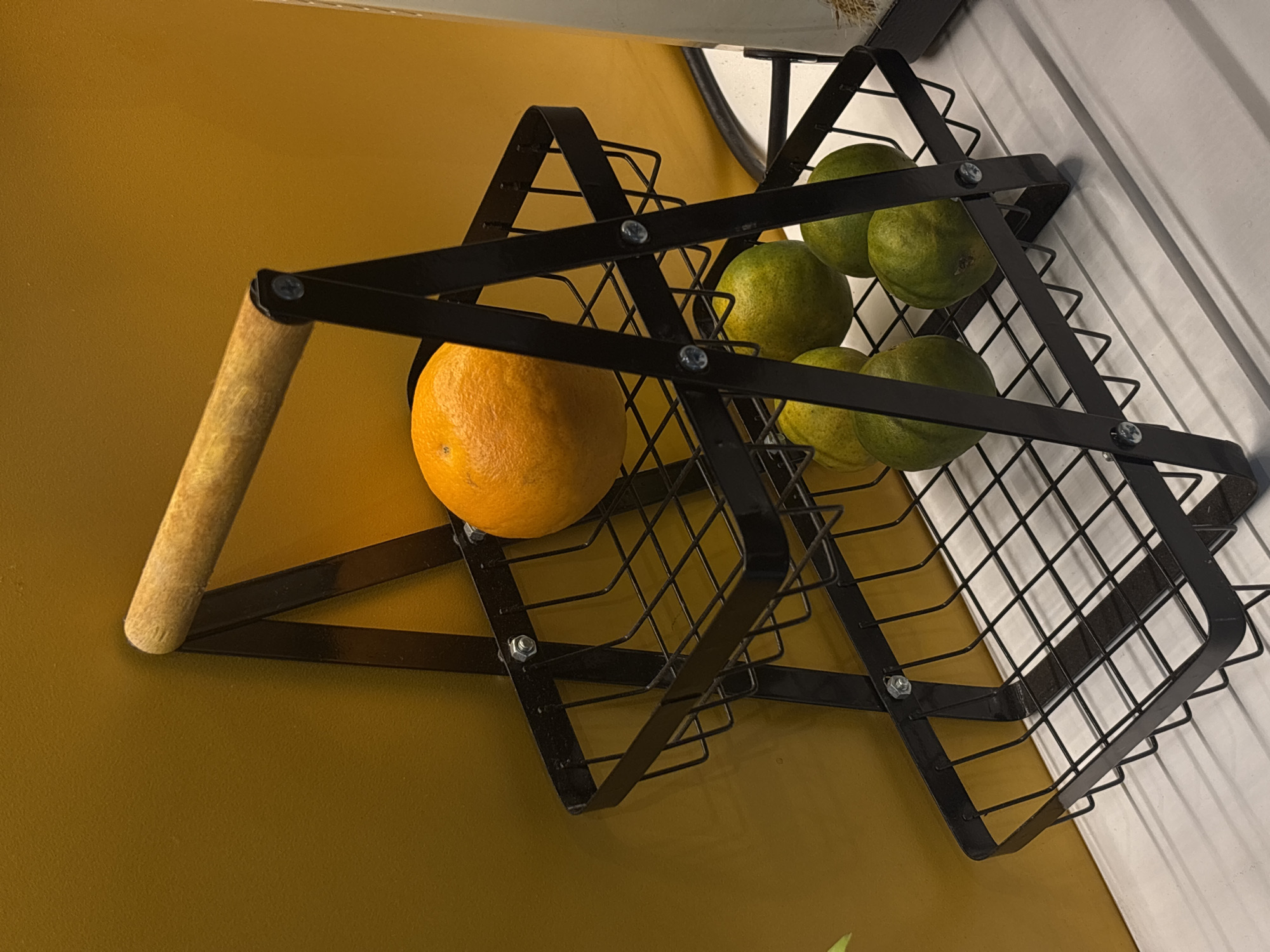 An indoor scene depicts a two-tiered black wire fruit basket with a wooden handle, positioned against a warm yellow wall. The upper basket contains a single, large orange. The lower basket holds several green citrus fruits, appearing to be limes or small oranges. The basket is supported by a black metal frame with visible screws. The surface beneath the basket appears to be a light-colored, slatted shelf or counter. There is no indication of the city of Semarang, Indonesia.