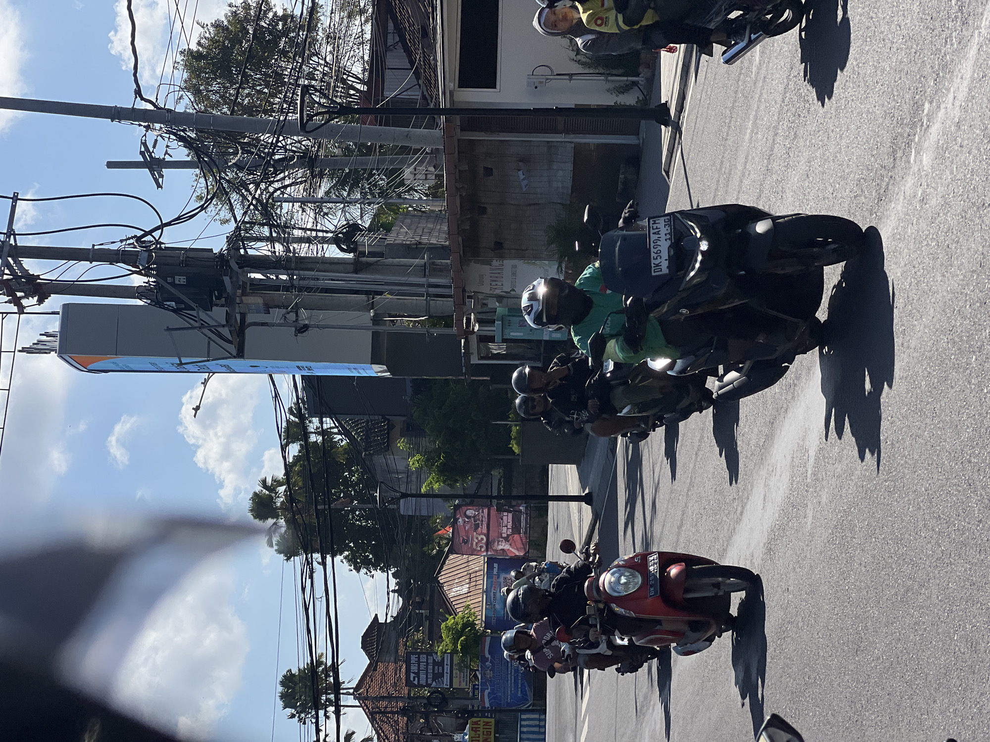 The image captures a street scene in Denpasar, Indonesia, on a sunny day with scattered clouds. Several motorbikes are being ridden in a single lane on an asphalt road. The foreground features a red scooter with a license plate visible and a black scooter with a rider wearing a green shirt and a helmet. To the right, another motorbike and rider are partially visible. In the background, there are buildings with traditional tiled roofs, utility poles with numerous wires, and some vegetation, including palm trees. Billboards and signs are also present on the buildings. The overall impression is one of daily urban life, with motorbike traffic being a prominent element.