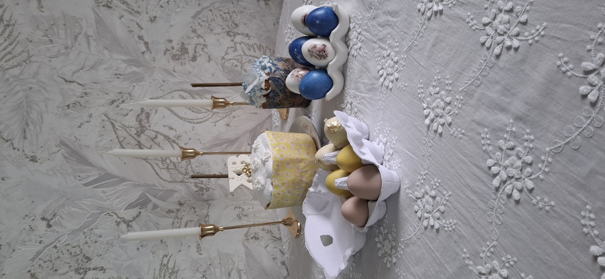 The image shows a table setting, likely for a celebration. There are two kulichi (Easter cakes) decorated with icing and edible decorations. One is covered in a yellow and white daisy-patterned paper, while the other has a blue and white floral pattern. There are also painted eggs in a decorative holder and in an egg carton. Three tall candlesticks with unlit white candles are present. The table is covered with a white embroidered tablecloth. The background is a patterned wallpaper. The image could have been taken in a private residence in Reshetnikovo, Russia.
