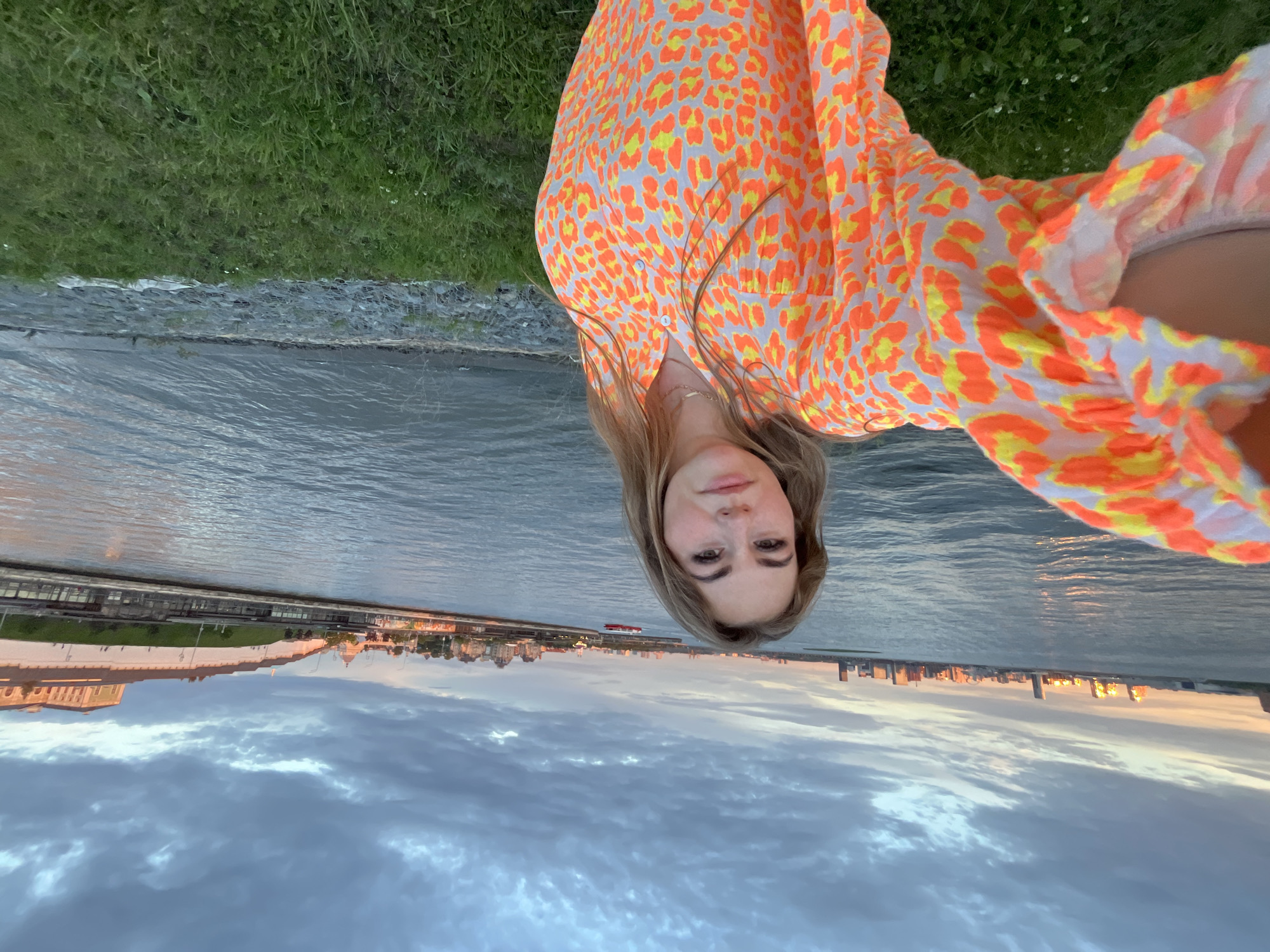 The media file shows a young woman with blonde hair, wearing a brightly colored orange and yellow animal print dress, standing near the water. The image is oriented sideways. She is facing the camera and has a neutral expression. In the background, there is a body of water, and beyond that, a cityscape with buildings and a cloudy sky. The sun appears to be setting, casting a warm glow on the city skyline. The scene suggests a relaxed atmosphere, possibly an evening stroll or a moment of reflection by the water. The location is identified as Kazan, Russia. No visible text is present in the image.