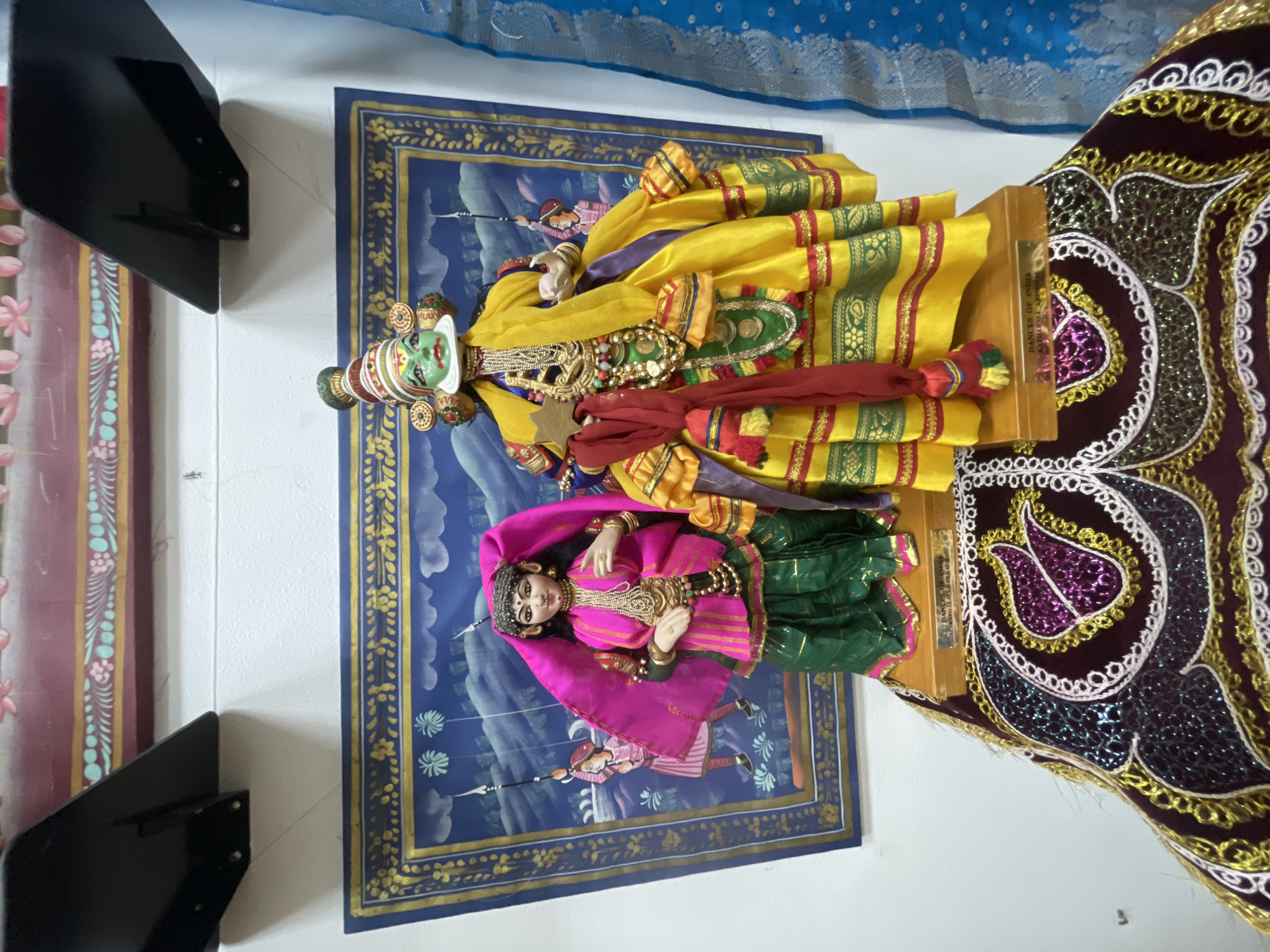 Two vibrantly dressed dolls, representative of Indian cultural figures, are centrally displayed in an indoor setting. The image is oriented with the dolls standing upright on a wooden base.
The prominent doll on the left is a male figure with a distinctive green face adorned with white and red markings, wearing a crown-like headpiece. He is elaborately dressed in a yellow and green layered garment, possibly a dhoti or traditional costume, accented with gold trim and red sashes. Intricate gold jewelry, including necklaces and armbands, decorates his body. He holds a small, light-colored object in his right hand.
To the right and slightly in front of the male doll is a female doll. She has fair skin, black hair, and delicate facial features, wearing a bright pink headscarf and a matching pink top with gold embroidery and beading. Her lower garment is a pleated green skirt, also embellished with gold. Gold jewelry adorns her neck and wrists.
The dolls stand on a wooden platform. On the front of this platform, text is visible: "DANCES OF INDIA" is clearly readable, followed by "KATHAKALI & BHARATANATYAM," indicating popular classical Indian dance forms.
Behind the dolls is a large blue tapestry or painting depicting a pastoral or mythological scene with two human figures holding spears, set against a backdrop of clouds and stylized vegetation. The tapestry has a gold border.
To the right of the dolls, a rich, dark maroon or purple fabric with elaborate gold, white, and iridescent purple embroidery featuring swirling patterns and paisley motifs is draped, possibly part of a larger textile display. In the upper background, a light blue fabric with a subtle textured pattern is visible. On the far left, a vertical panel with a pink-red and light blue floral pattern contributes to the decorative environment. A black, angular object, possibly a shelf or bracket, is positioned in the foreground on the left.
The overall scene suggests a curated display of Indian cultural artifacts, possibly for sale or exhibition, with a focus on traditional dress and performing arts, presented in Sochi, Russia.