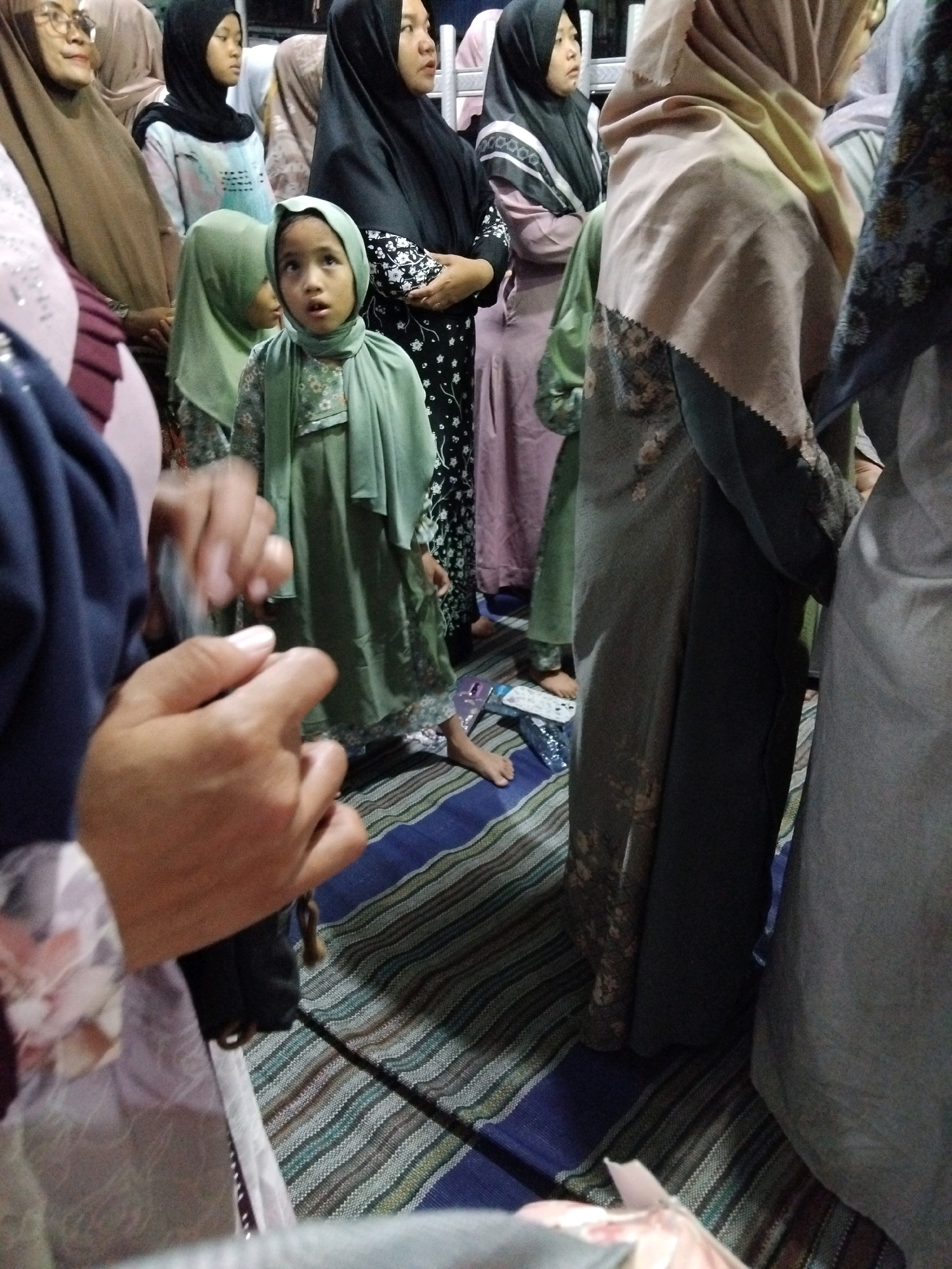The image depicts a group of people, primarily women and a young child, gathered indoors. They are wearing traditional Islamic clothing, including various colored hijabs and long garments. The child in the foreground, wearing a green hijab and dress, appears to be looking upwards with a surprised expression. The individuals are standing on a patterned rug. The scene suggests a casual gathering or event. The location is identified as Kepuh Kiriman, Indonesia.