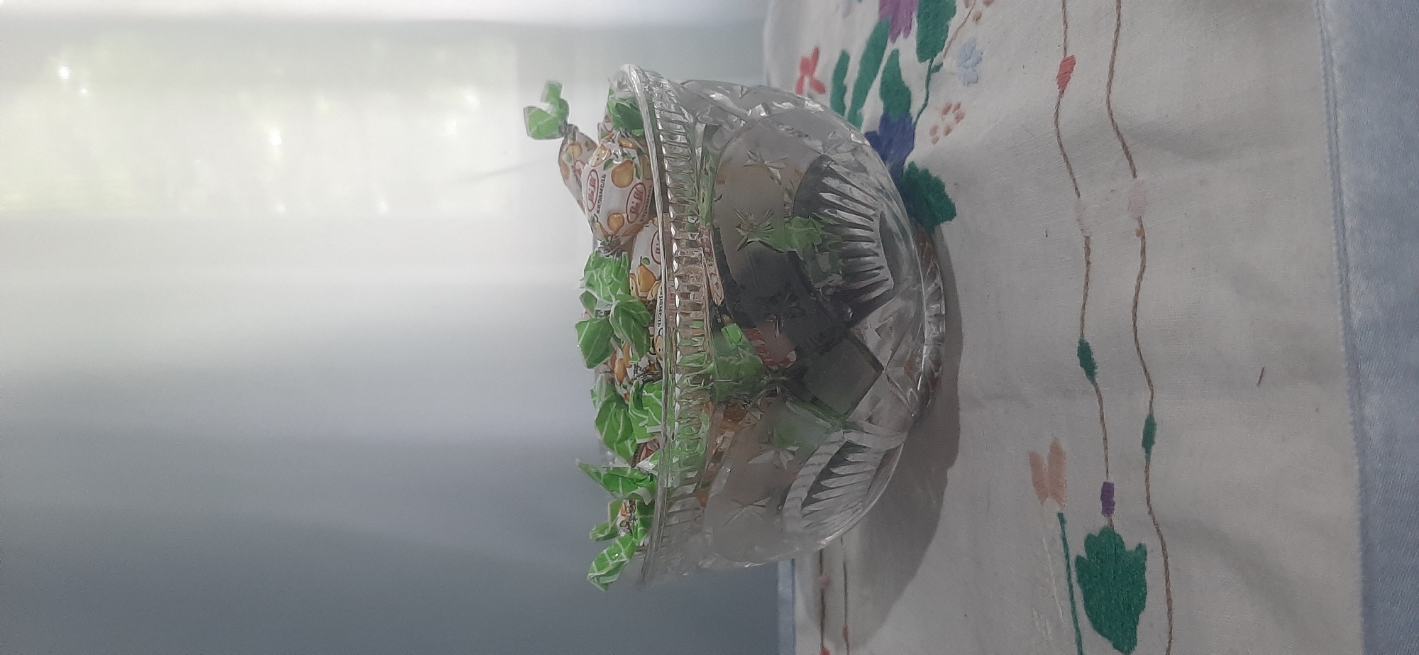 The image shows a crystal bowl filled with individually wrapped candies. The bowl is intricately cut with star and diamond patterns. The candies are wrapped in green and white foil, with some wrappers featuring a drawing of oranges and text in a Cyrillic script. The bowl is placed on a white tablecloth with a delicate embroidered floral pattern in shades of green, pink, and purple. The background is out of focus, with a bright, diffused light source, suggesting an indoor setting with ample natural light. The scene is static, with no discernible activity other than the display of the candies. The overall impression is one of simple domesticity and perhaps a sweet treat offered to guests. The text on the candy wrappers is partially visible, and appears to be in Russian, consistent with the location context of Yessentukskaya, Russia. No people are visible in the frame, and the time of day and weather are not clearly indicated, though the bright light suggests daytime.