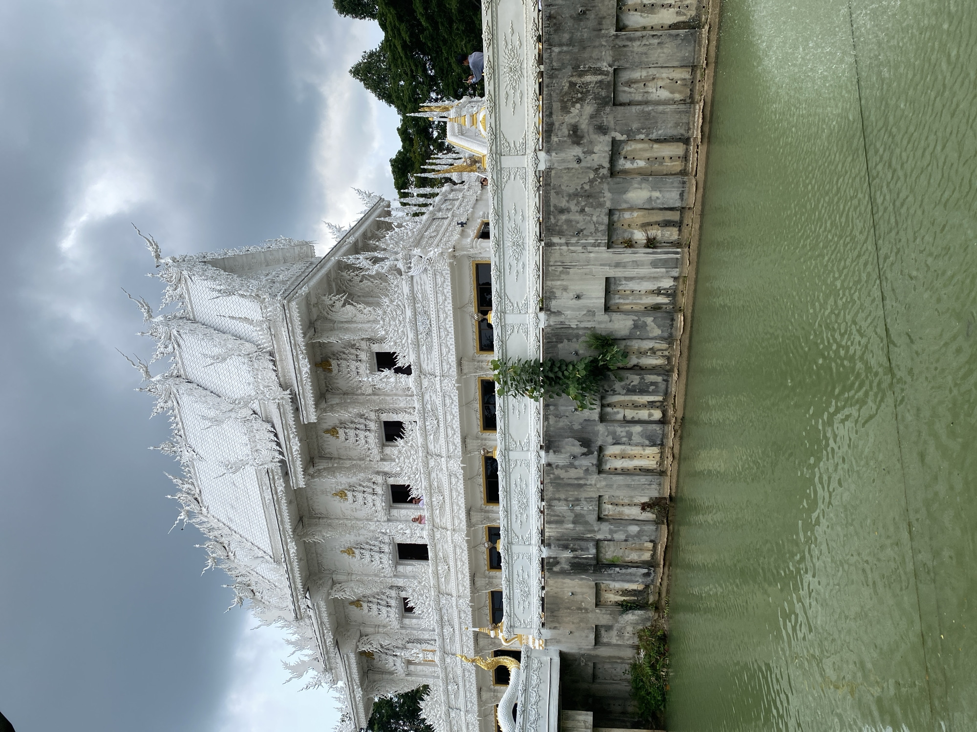 The image shows a building with ornate white facades, potentially a temple or religious structure, reflected in a murky green body of water. The building has multiple levels, with windows and decorative elements. The sky is overcast and stormy. A section of concrete with rectangular indentations is visible alongside the water, and some greenery grows between the water and the building. The location is likely in Thep Krasatti, Thailand, given the cultural style of the building. A person in a pink shirt is visible behind the windows in the building.