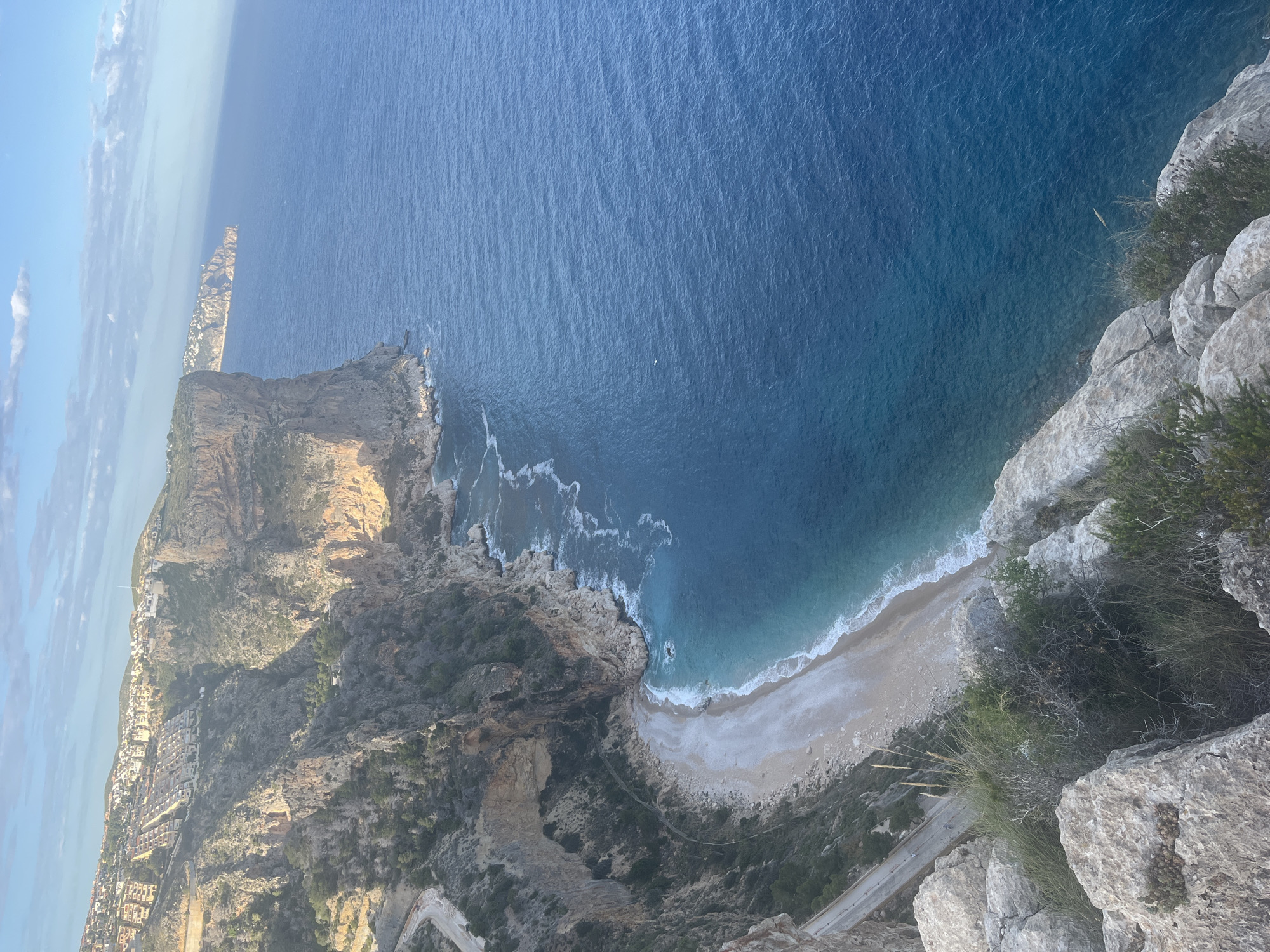 The image shows an aerial view of Cala del Moraig in El Poble Nou de Benitatxell / Benitachell, Spain. A steep, rocky cliff dominates the foreground on the right side of the image, with scattered vegetation growing on its surface. Below the cliff is a small, white pebble beach where waves gently break along the shoreline. The turquoise water transitions to a deeper blue color as it stretches further into the distance. Another rocky cliff is visible in the background on the left side of the image, with buildings and structures of the town visible along the top of the cliff. A winding road is seen snaking down the cliffside towards the beach.