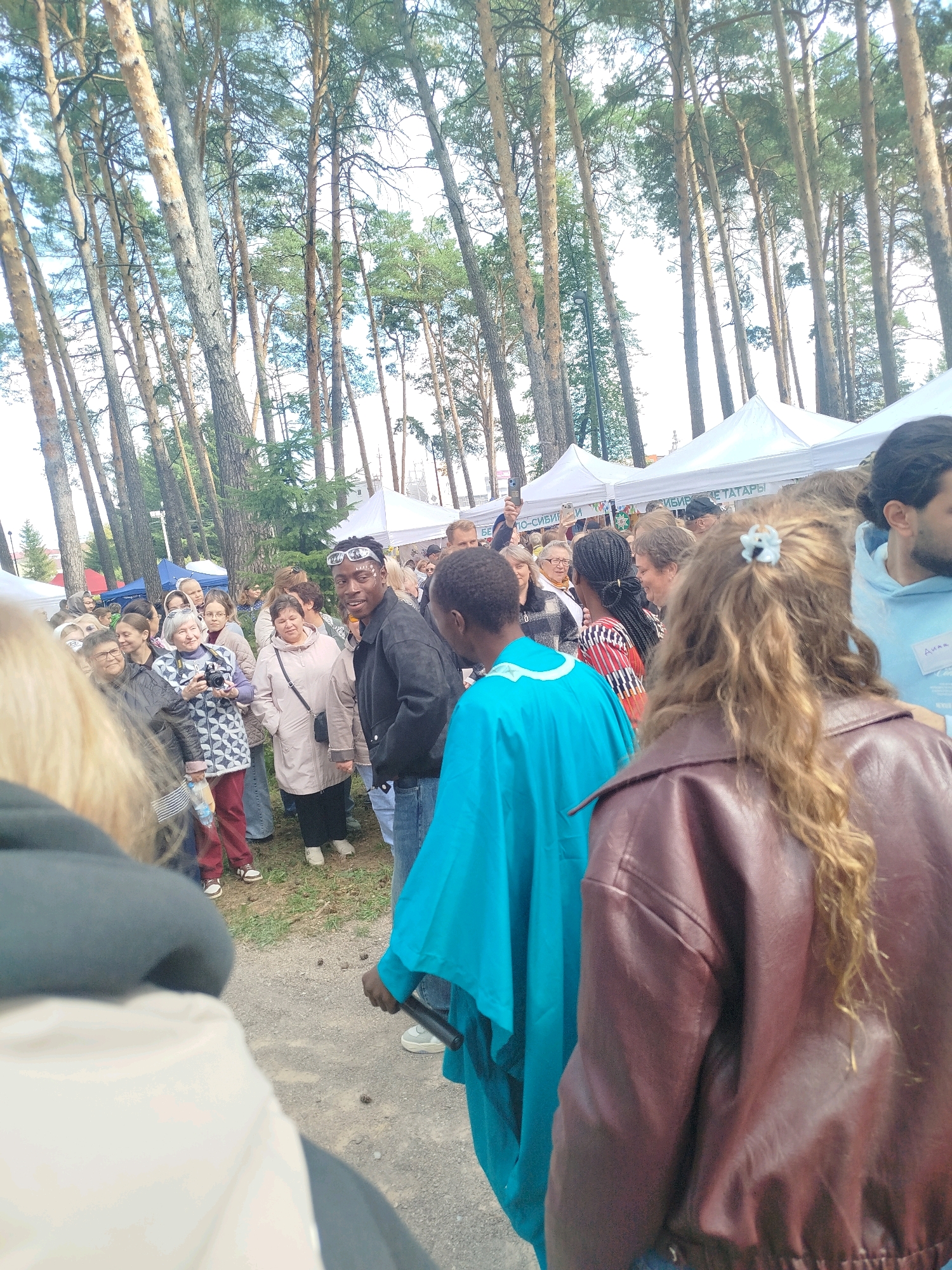 The image depicts an outdoor gathering in a wooded area, likely a festival or market, with white tents visible in the background. The scene is populated by a diverse group of people of various ages and ethnicities, engaged in conversation and observation. Some individuals are dressed in casual attire, while others wear more distinctive clothing, such as a man in a bright turquoise robe. The environment is characterized by tall pine trees and a dusty ground. The presence of tents suggests a temporary setup for an event. The overall atmosphere appears lively and social. The provided crops help to focus on specific individuals and details within the larger scene, revealing facial expressions, clothing textures, and the presence of objects like a camera and a baton. The specific location within Zavodoukovsk, Russia, cannot be determined from the image alone but the context suggests an event being held in a park or recreational area.