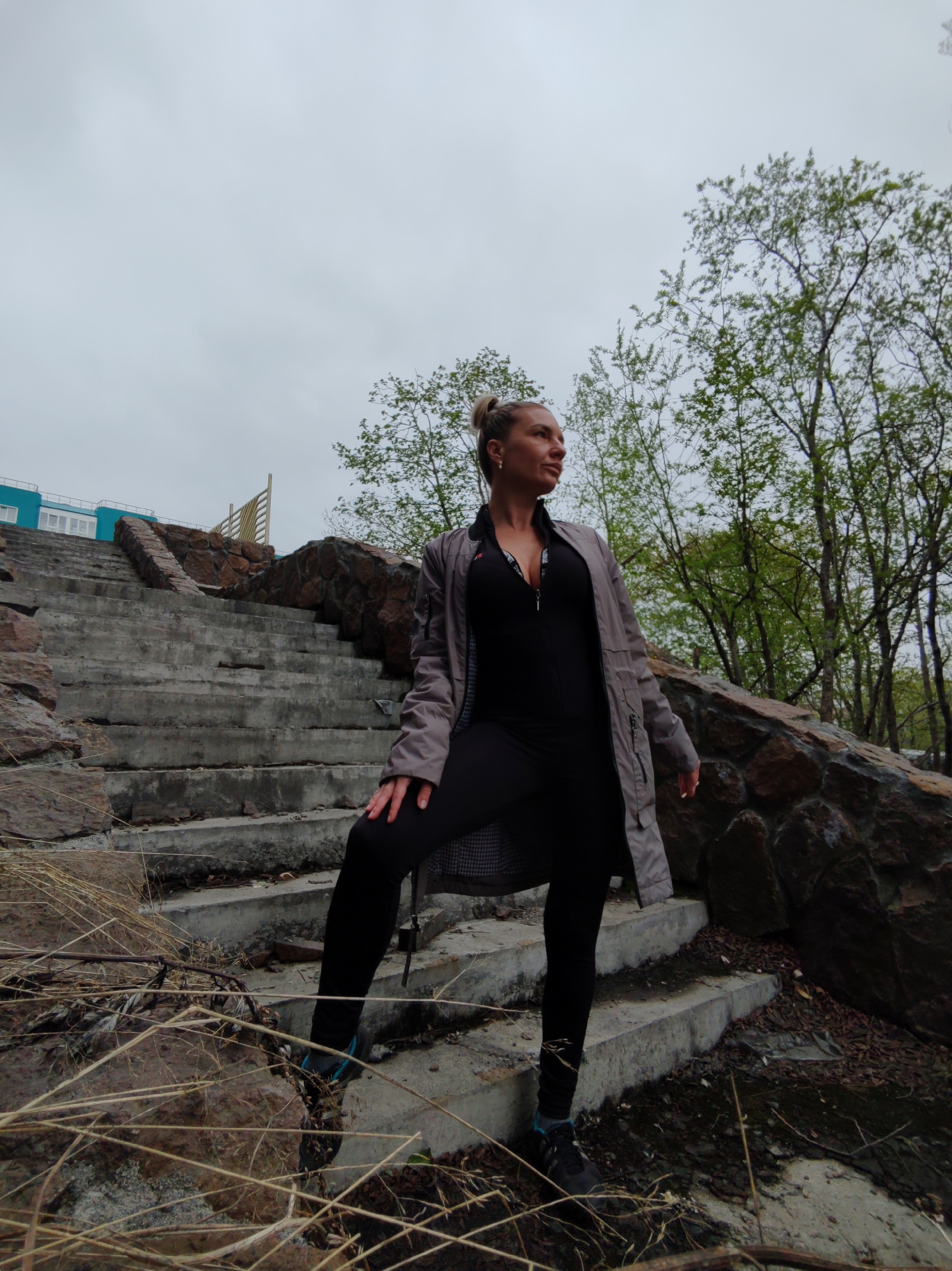 A woman stands on a concrete stairway with stone retaining walls on either side. The stairway leads up towards a blue building with white windows. Trees with green leaves are visible behind the woman and the stairway. The sky is overcast. The woman is wearing a black jumpsuit, a grey jacket, and black pants. She has her hair tied up in a bun. The image appears to have been taken in Petropavlovsk-Kamchatsky, Russia, as indicated by the location.