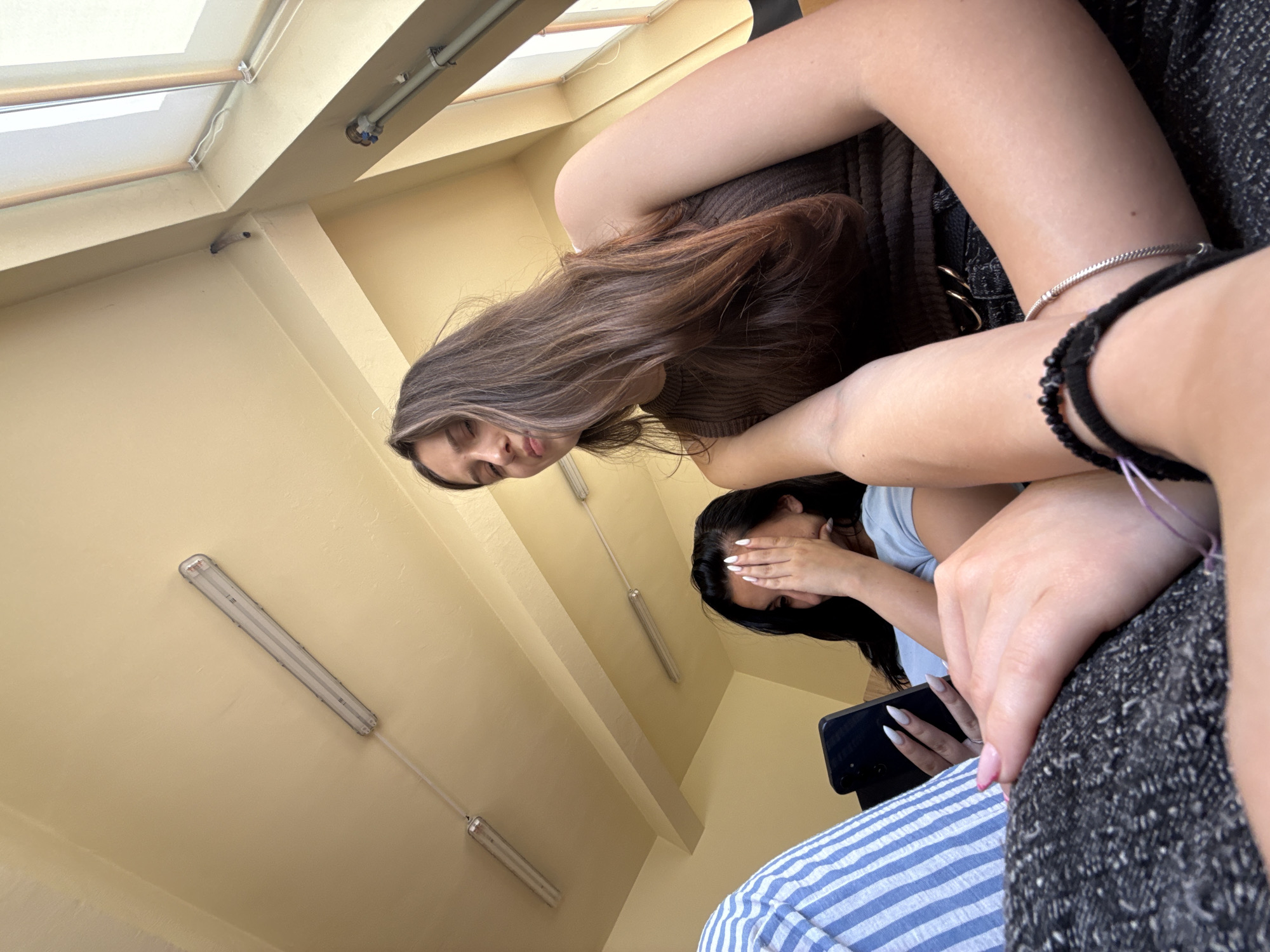 The image shows two young women in a classroom setting. The woman in the foreground has long brown hair and is wearing a brown sleeveless top and dark pants. She is looking down at her phone. The woman behind her has dark hair and is covering her face with her hand. She is also looking at her phone. The classroom has a yellow ceiling with fluorescent lights and a window with blinds. The image appears to be taken from a low angle, looking up towards the ceiling and window. The location is identified as Sofia, Bulgaria.