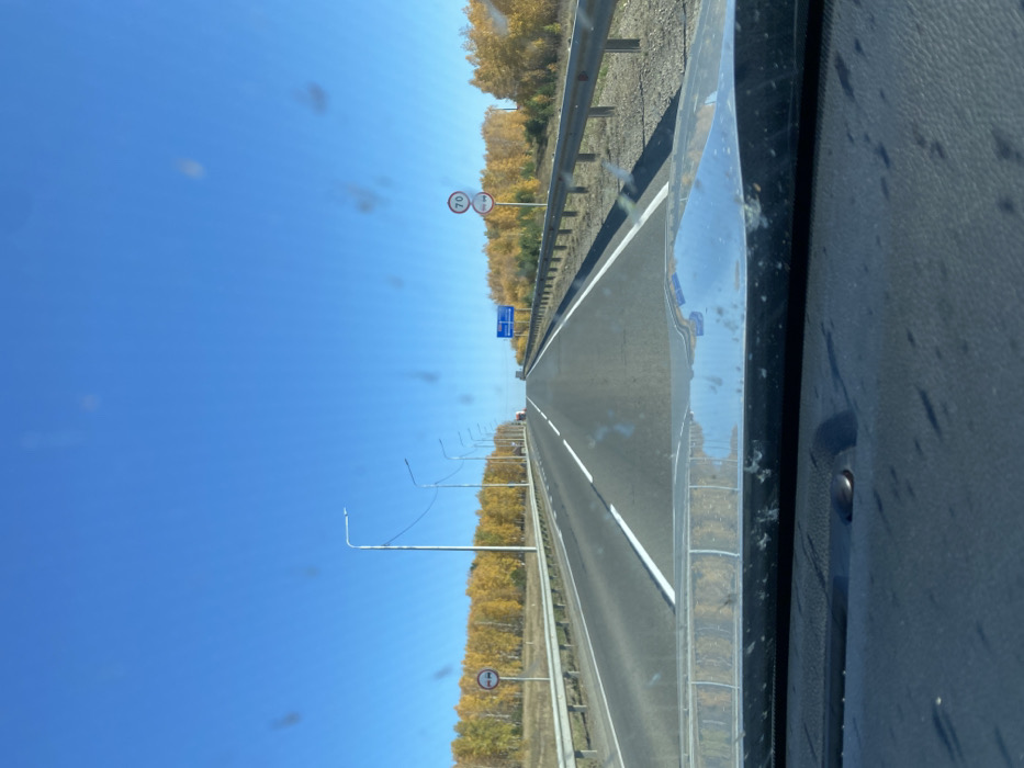 This image captures a multi-lane highway near Kuytun, Russia, on a clear autumn day, viewed from the interior of a vehicle. The foreground shows the dark-colored, textured dashboard and a reflective chrome-like strip, which mirrors the blue sky and distant foliage. The windshield, through which the scene is observed, exhibits several circular dust or smudge marks.
The asphalt highway stretches directly ahead, presenting two visible lanes marked by dashed white lines, bordered by a solid white line on the right shoulder. Metal guardrails line both sides of the road. On the left, a series of evenly spaced streetlights with slim poles recedes into the distance. A circular white sign with a red border on the left side depicts two red vehicle silhouettes—a passenger car and a lorry—positioned side-by-side within the circle. Beyond the guardrail, a dense stand of trees displays vivid yellow and orange autumn foliage.
On the right side of the road, two stacked circular signs are present. The upper sign is white with a red border and displays "70" in black, indicating a 70 km/h speed limit. The lower sign is a blue circle with a red border and a single red diagonal line, signifying "No Stopping" (Russian sign 3.27). Further along the highway, a large blue rectangular informational sign with white text is mounted on an overhead structure. A similar line of bright autumn trees borders the right side of the road.
In the far distance, several vehicles, including what appears to be a large truck, are visible on an ascending section of the highway. The sky is uniformly brilliant blue and entirely cloudless, indicating direct sunlight conditions.
