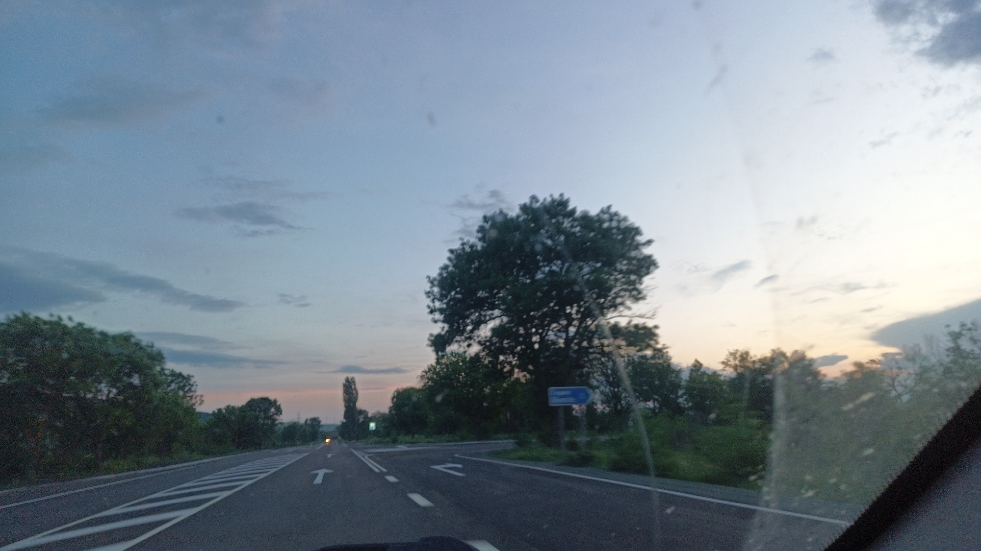The image shows a view from inside a car on a road leading into the city of Glumche, Bulgaria. The road has white dashed lines marking lanes and arrows indicating directions. A T-intersection is visible with a blue sign pointing toward Glumche. The sky is overcast, with hues of pink and blue indicating either sunrise or sunset. Trees and greenery line the sides of the road. A few other vehicles are visible in the distance. The foreground includes part of the car's dashboard and windshield, with some smudges on the glass.